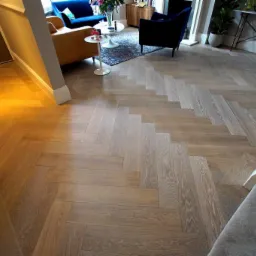 Ułożona w jodełkę jasna podłoga z paneli drewnianych przechodząca z przedpokoju do salonu z niebieską sofą i fotelami.