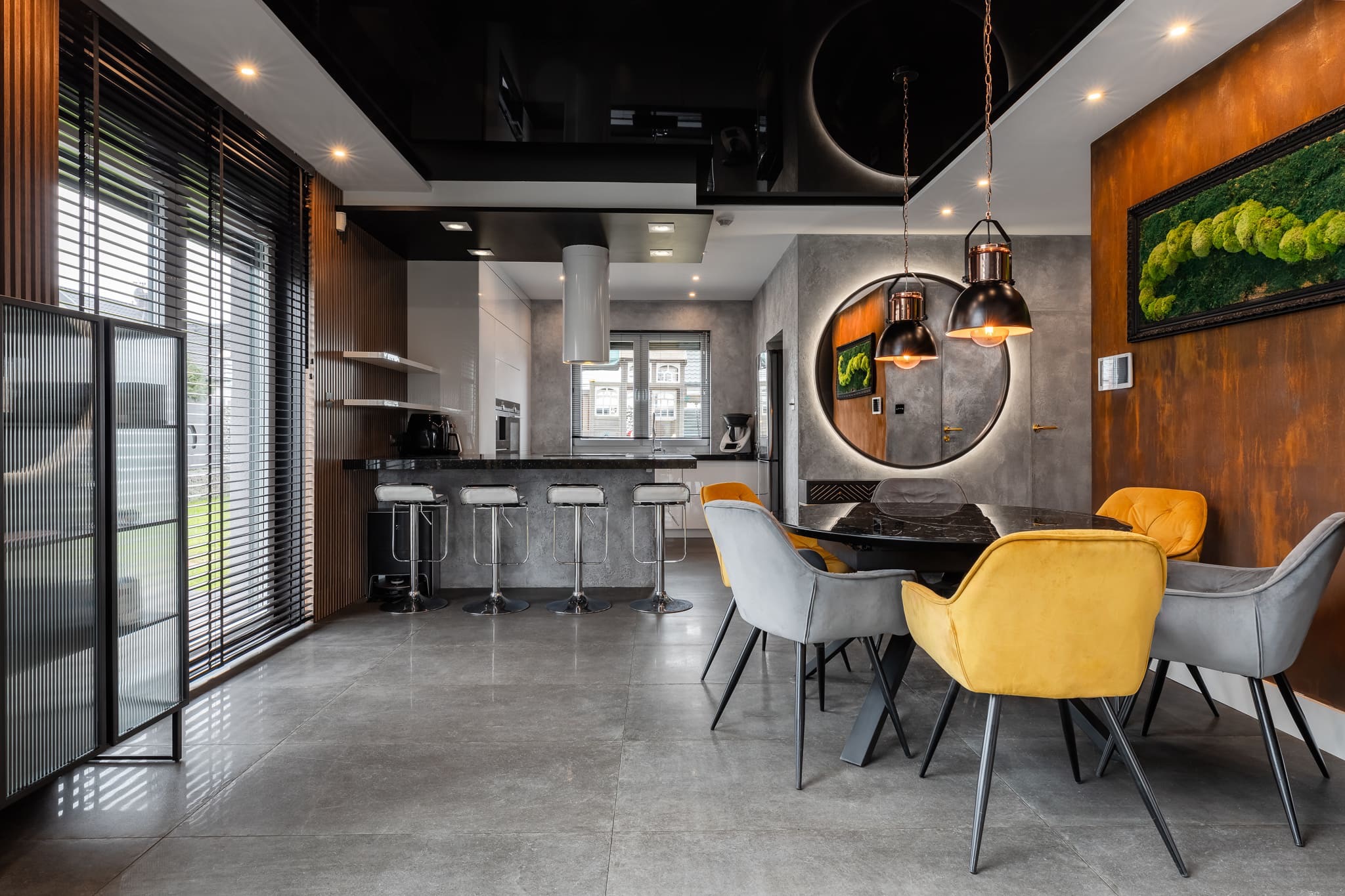 Nowoczesne wnętrze: szara podłoga, czarny sufit z lustrzanym połyskiem, wyspa kuchenna z hokerami i okrągłe lustro nad stołem z krzesłami w odcieniach szarości i żółci.