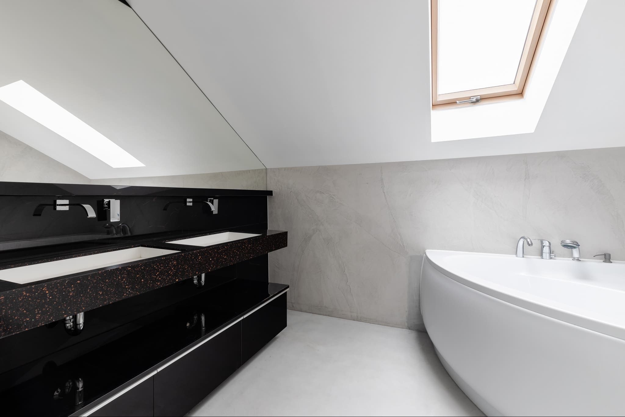 Nowoczesna łazienka na poddaszu z dwoma umywalkami na czarnym blacie z brokatem i białą wanną, oświetlona oknem dachowym.