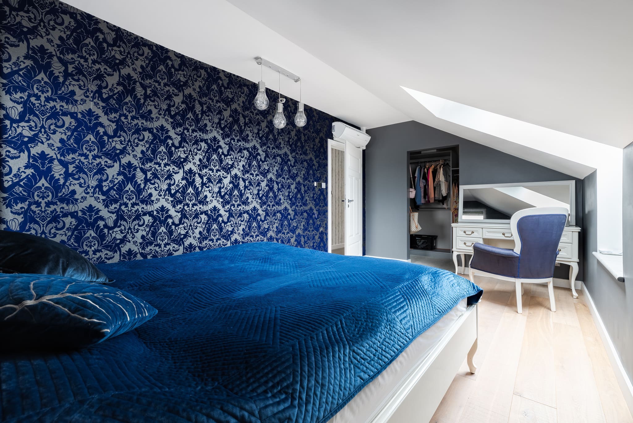 Sypialnia na poddaszu z tapetą w niebieskie wzory, łóżkiem z aksamitną narzutą i garderobą. Charakterystyczne skosy i oświetlenie.