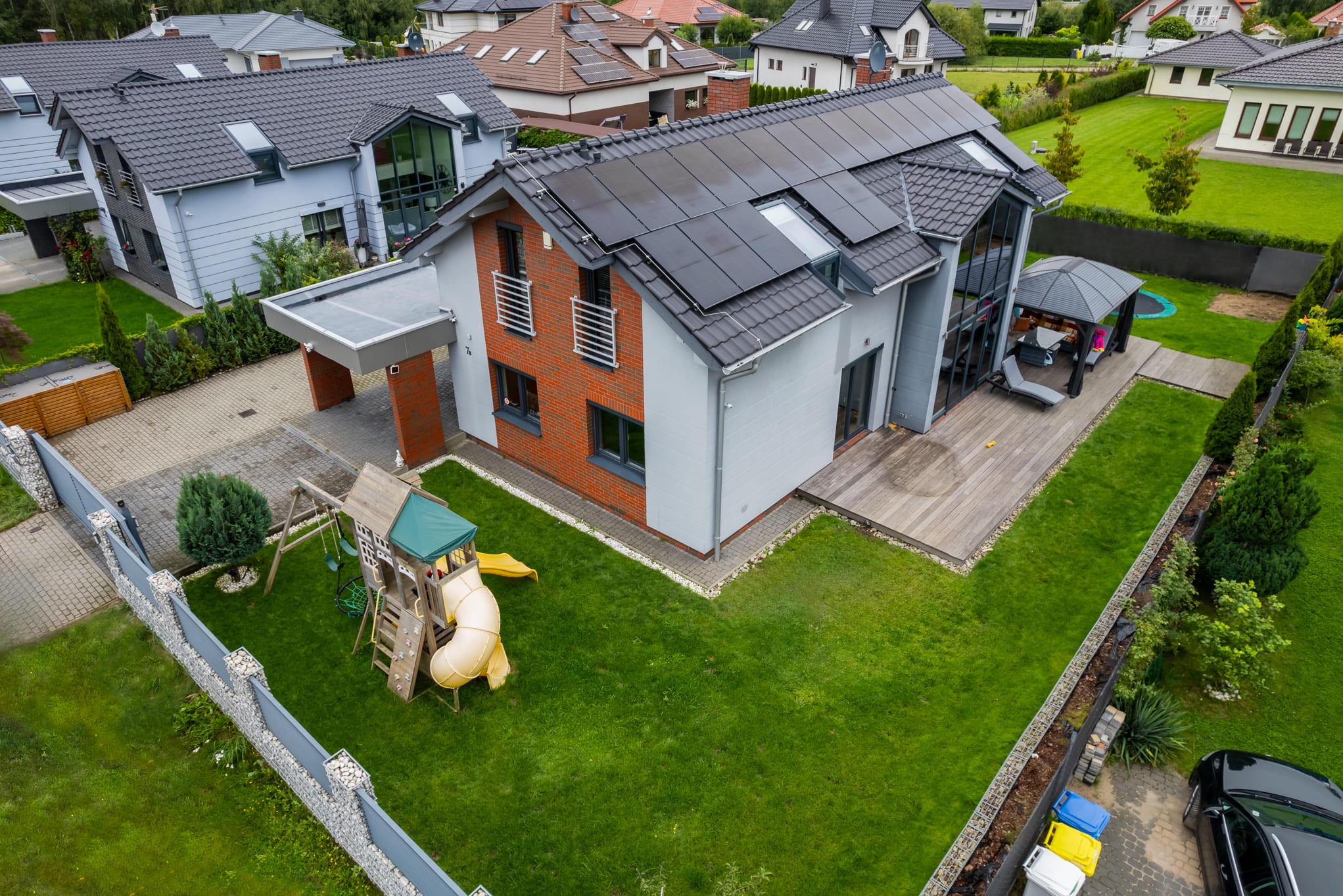 Nowoczesny dom z panelami słonecznymi na dachu, widok z góry. Na posesji plac zabaw i altana. Elementy ceglanej elewacji, szara kostka brukowa i zadbany trawnik.