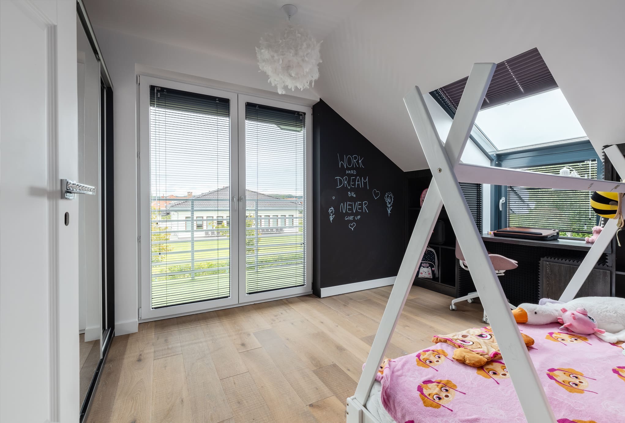 Jasne wnętrze pokoju dziecięcego na poddaszu z drewnianą podłogą, oknami z żaluzjami i ścianą pomalowaną farbą tablicową z napisem.