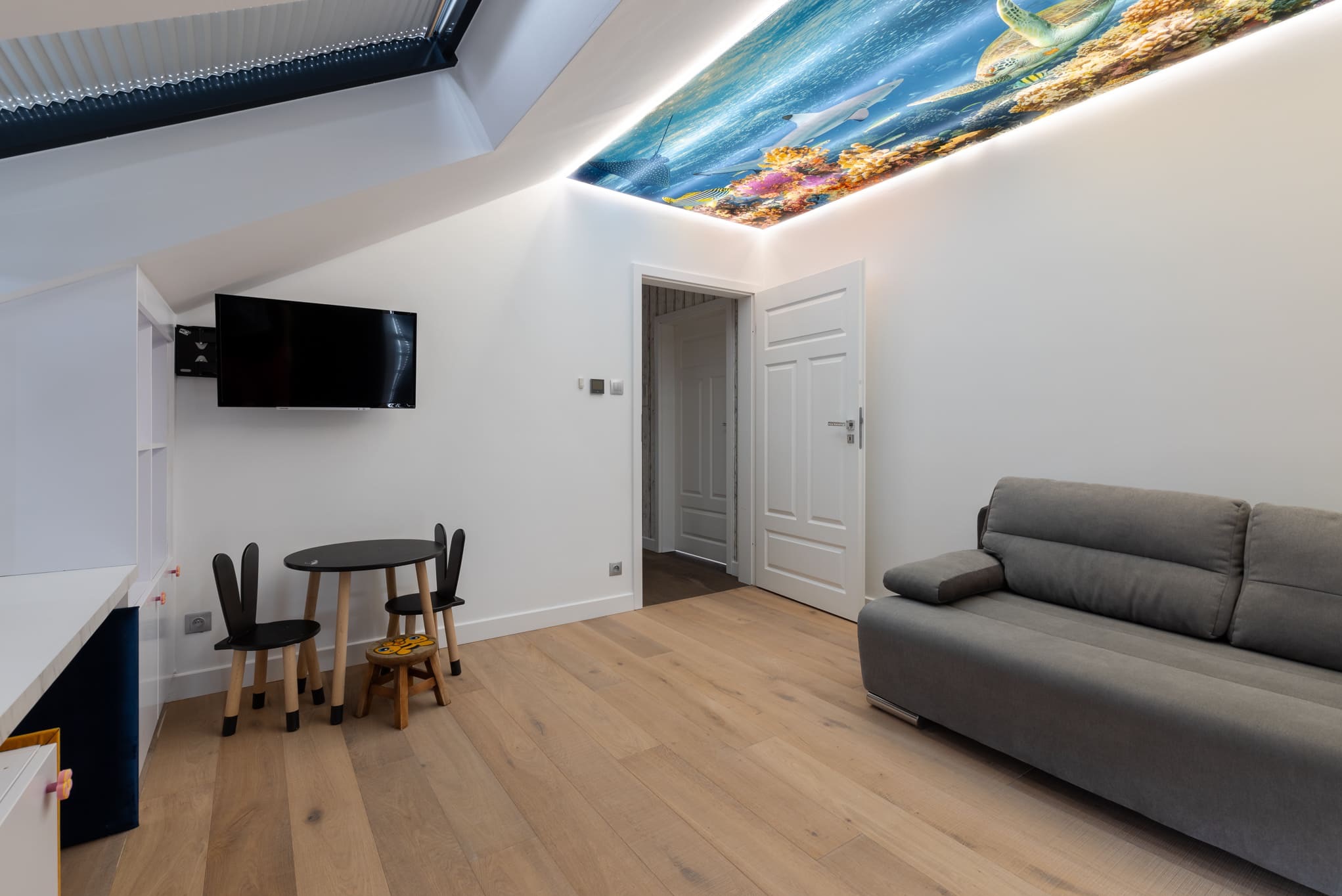 Pokój dziecięcy z podświetlanym sufitem napinanym z motywem podwodnego świata. Widoczny szary narożnik, mały stolik i telewizor na ścianie.