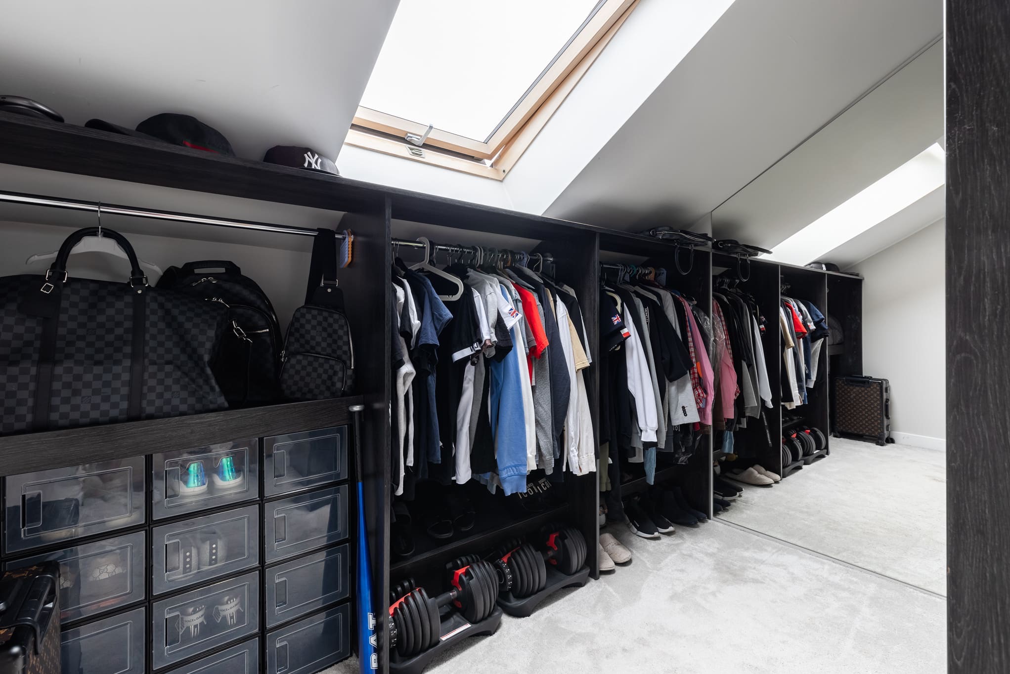 Przestronna garderoba z ciemnego drewna, oświetlona światłem z okna dachowego. Ubrania wiszą na wieszakach, a na półkach leżą torby i akcesoria sportowe.