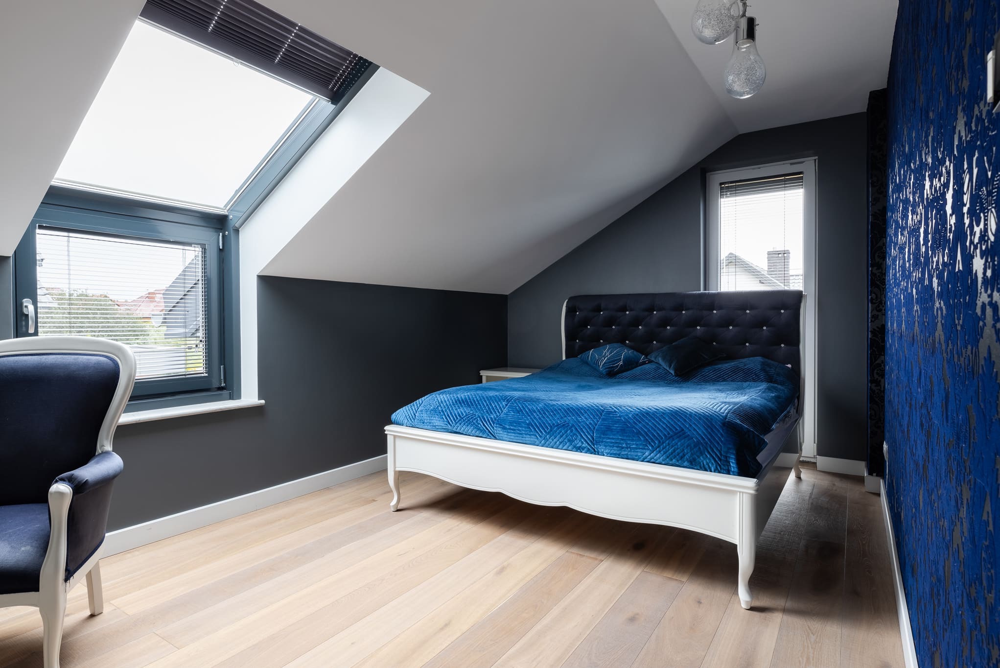 Sypialnia na poddaszu z białym łóżkiem z niebieską narzutą, oknami dachowymi z żaluzjami i ciemnymi ścianami z wzorzystą tapetą. Widoczne drewniane panele podłogowe.