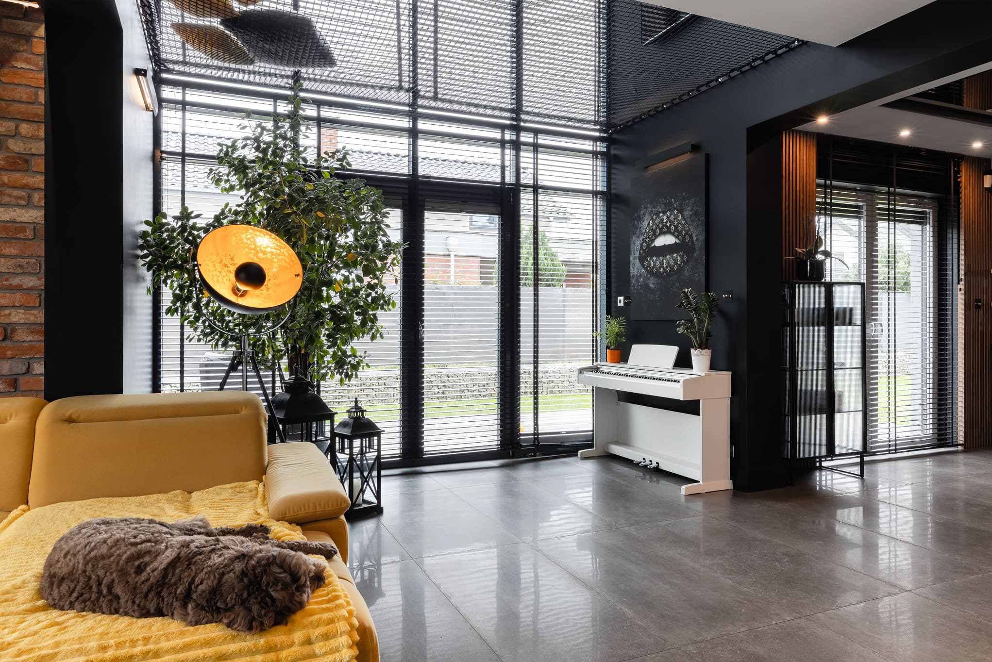Nowoczesny salon z szarymi płytkami, kanapą i psem. Duże okna z żaluzjami, białe pianino i lampa stojąca tworzą elegancki wystrój wnętrza.