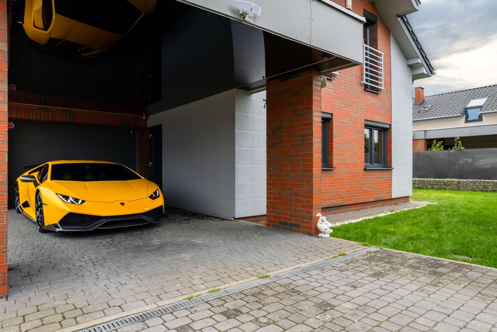 Żółte Lamborghini w garażu nowoczesnego domu z ceglaną elewacją i kostką brukową. Widoczna figura kaczki przy budynku i fragment drugiego auta w garażu.