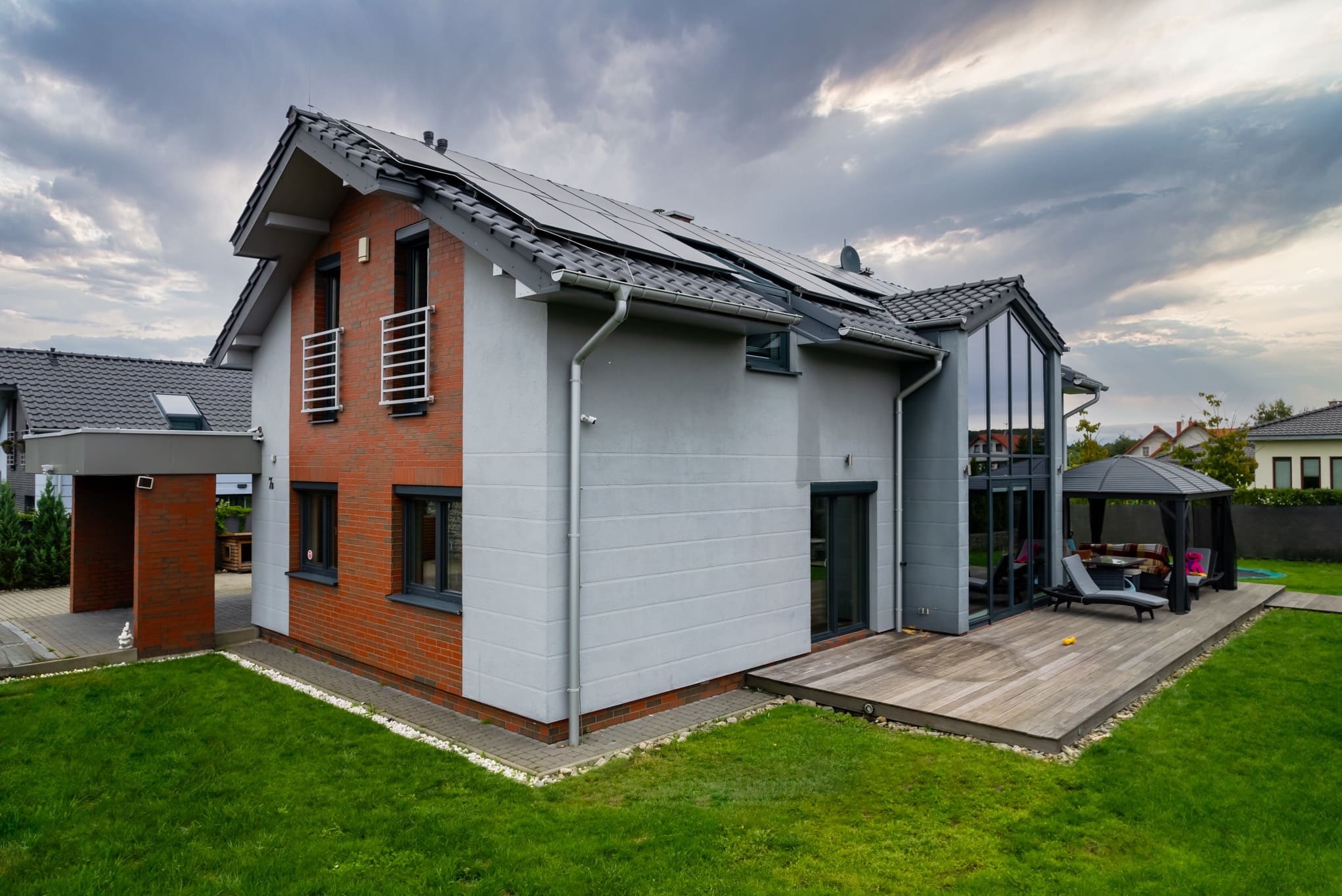 Nowoczesny dom z panelami słonecznymi na dachu, drewnianym tarasem i elementami cegły. Widok z zewnątrz na zadbany ogród i altanę.