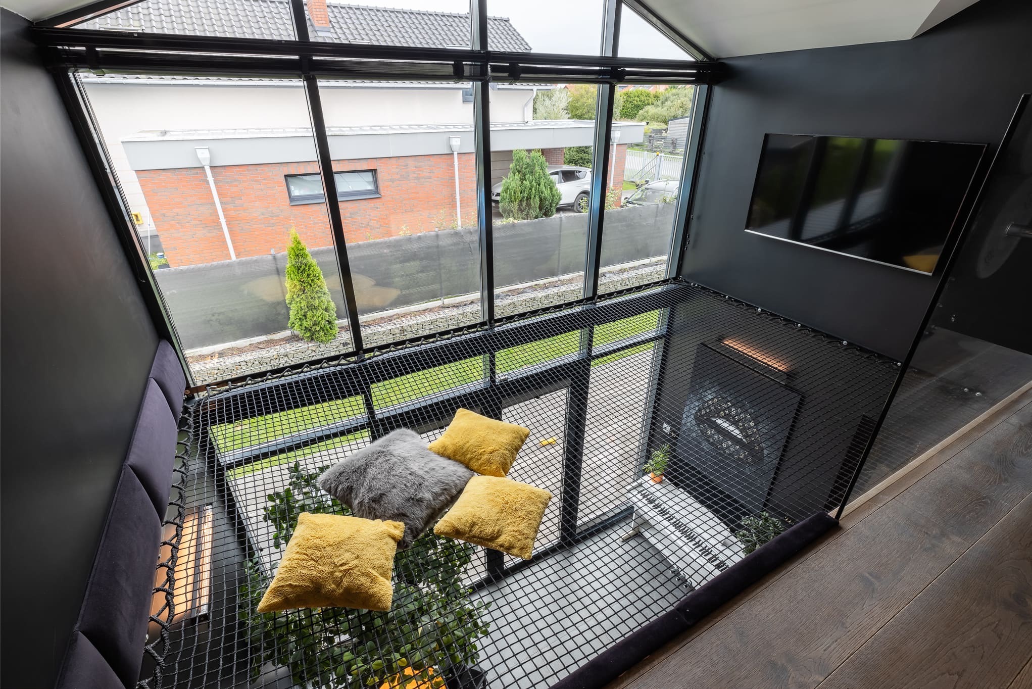 Nowoczesne wnętrze z siatką relaksacyjną pod sufitem, poduszki dekoracyjne, duże okna z widokiem na zewnątrz i czarny telewizor na ścianie. Nietypowy element wystroju.