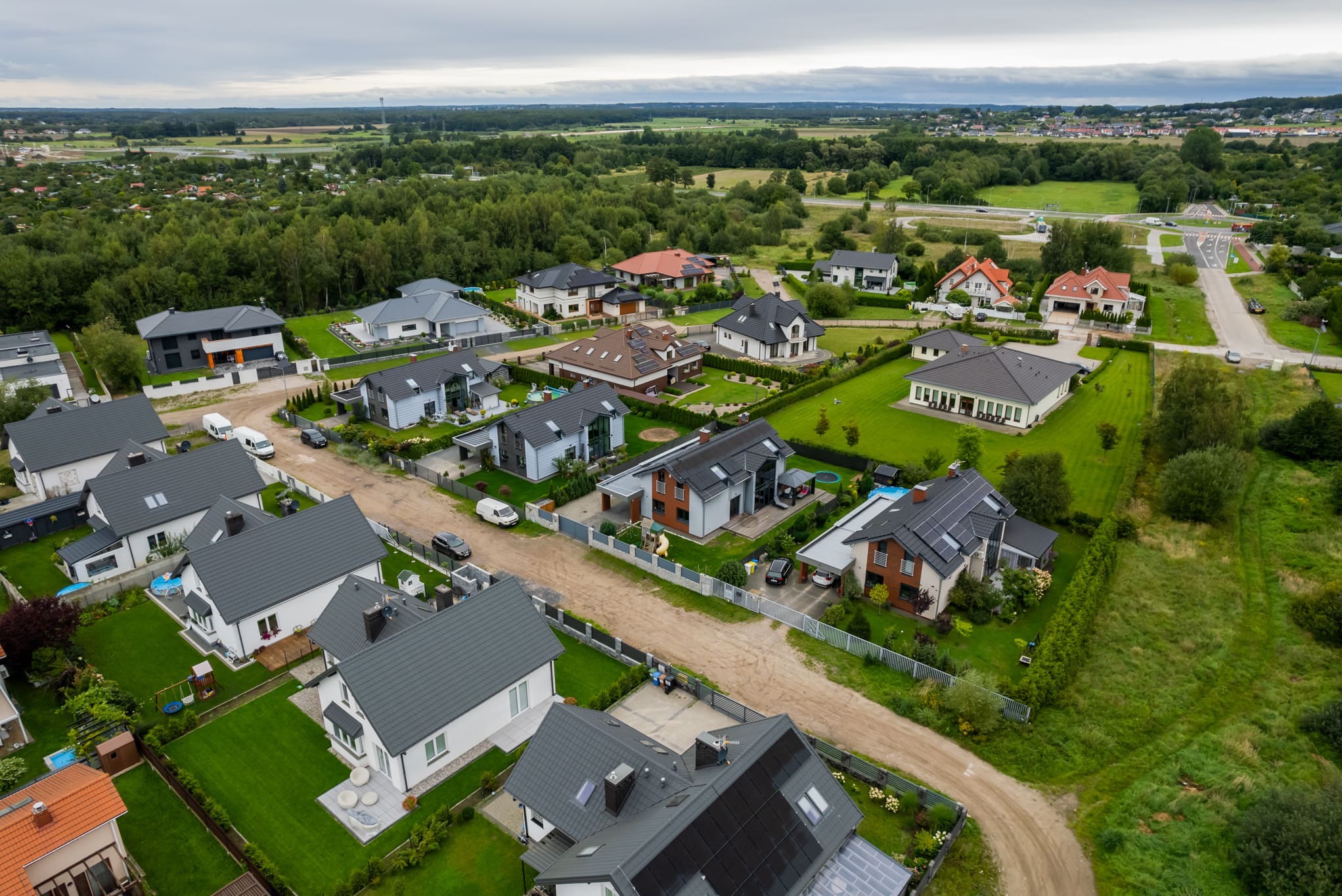 Panoramiczny widok z lotu ptaka na nowoczesne domy jednorodzinne z szarymi dachami i panelami słonecznymi, otoczone zielenią i polami w słoneczny dzień.