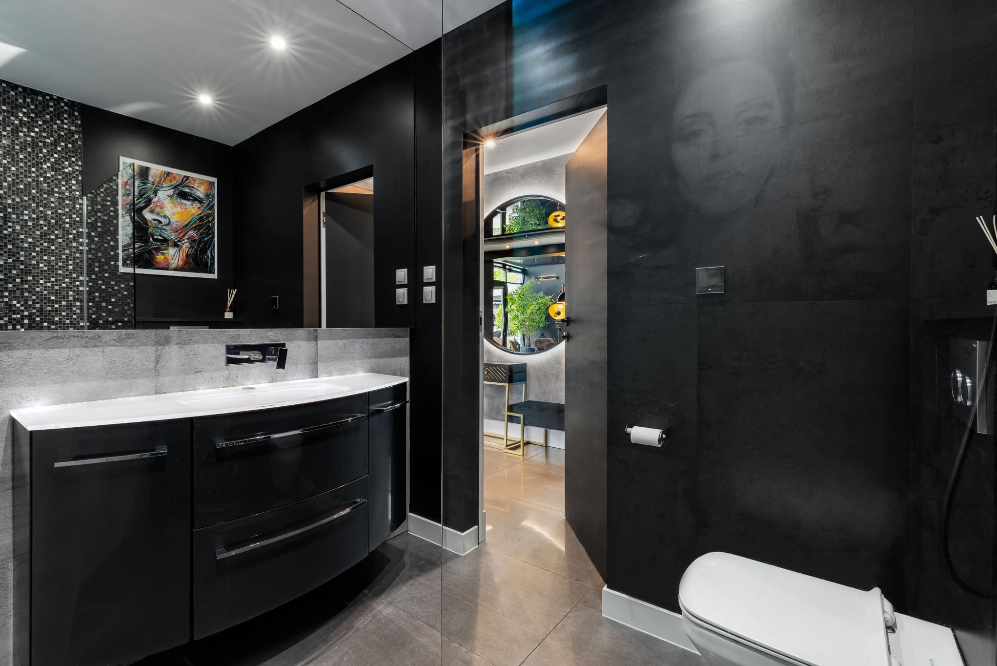 Czarne, eleganckie wnętrze łazienki z umywalką, lustrem i dekoracyjną grafiką. W tle widoczne kolejne pomieszczenie z okrągłym lustrem i roślinami.
