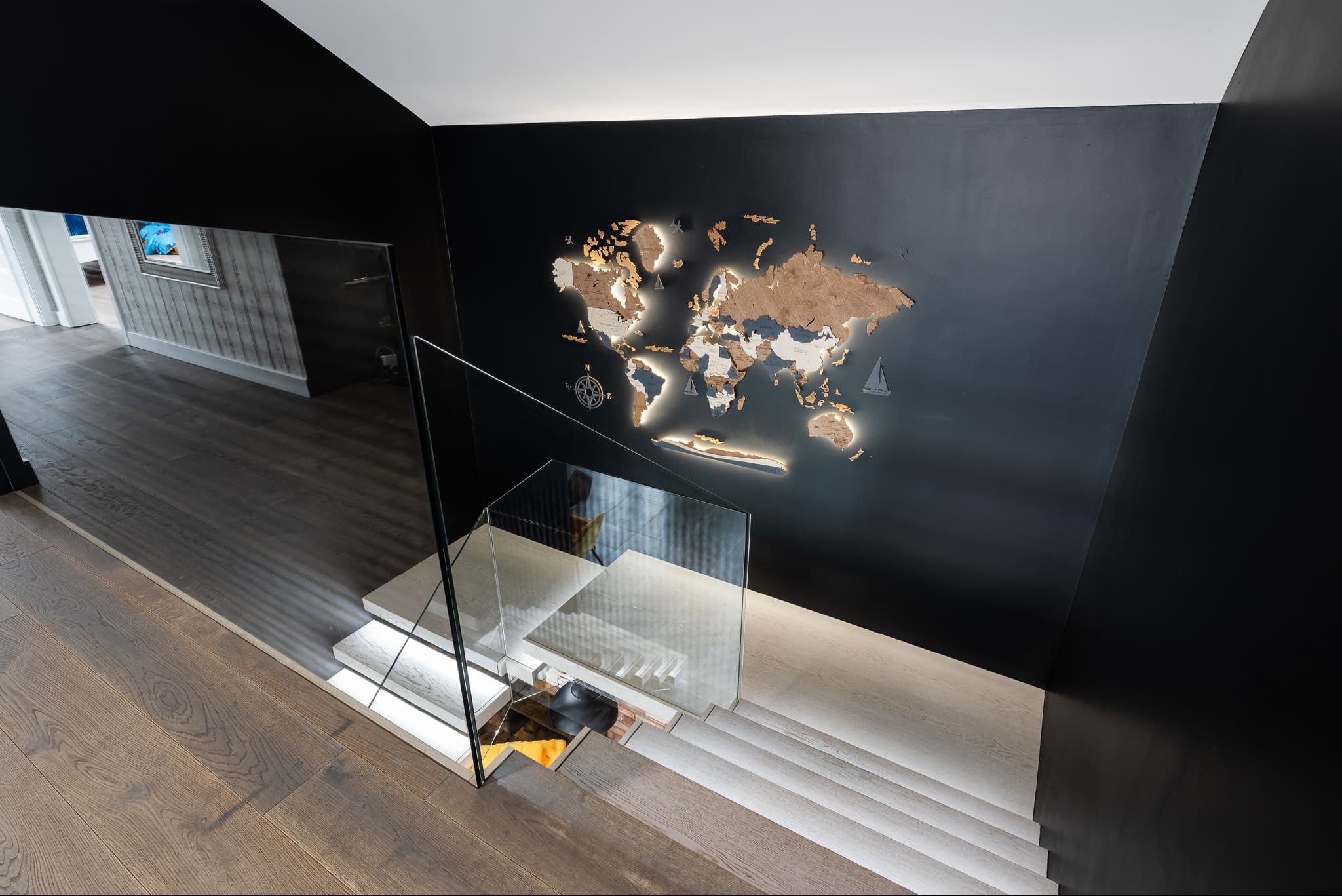 Nowoczesne schody z drewnianymi stopniami i szklaną balustradą. Na czarnej ścianie podświetlana drewniana mapa świata z motywami żeglarskimi.