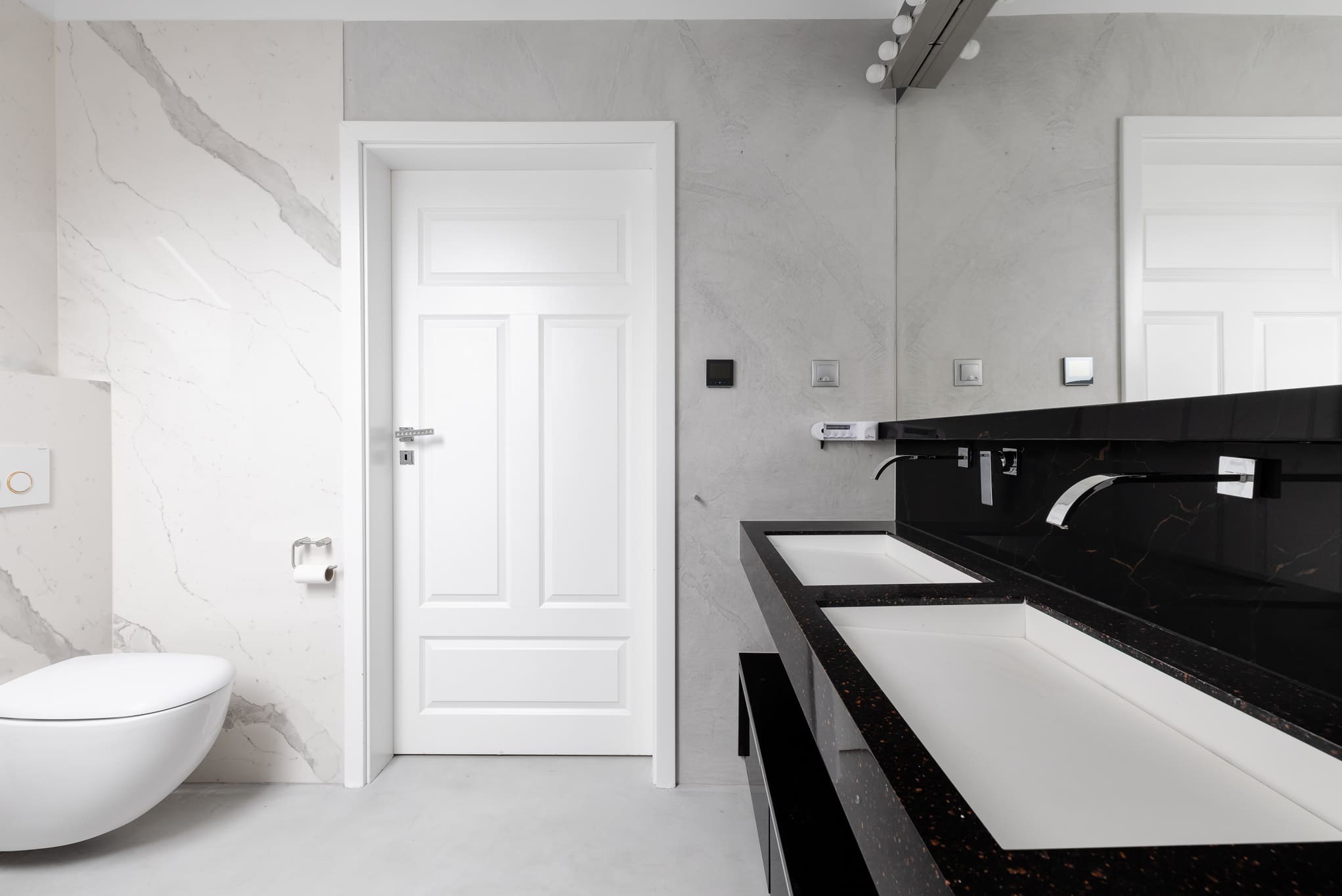 Nowoczesna łazienka w bieli i szarości z czarnym blatem umywalki. Minimalistyczny design, marmurowe ściany, podwieszana toaleta i oświetlenie.