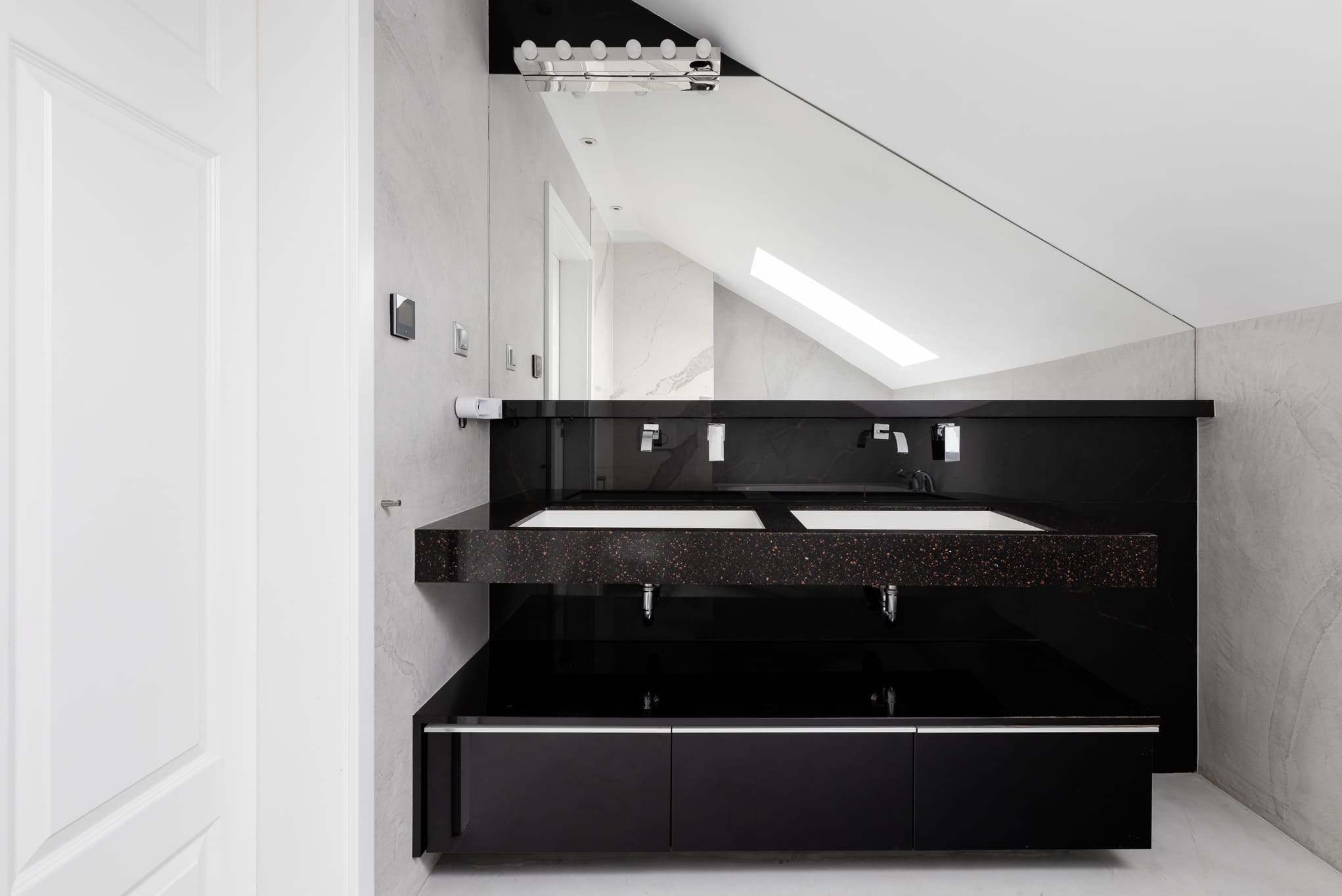 Czarne, podwójne umywalki z blatem w łazience na poddaszu, z lustrem sięgającym sufitu i białymi drzwiami po lewej. Minimalistyczny design.
