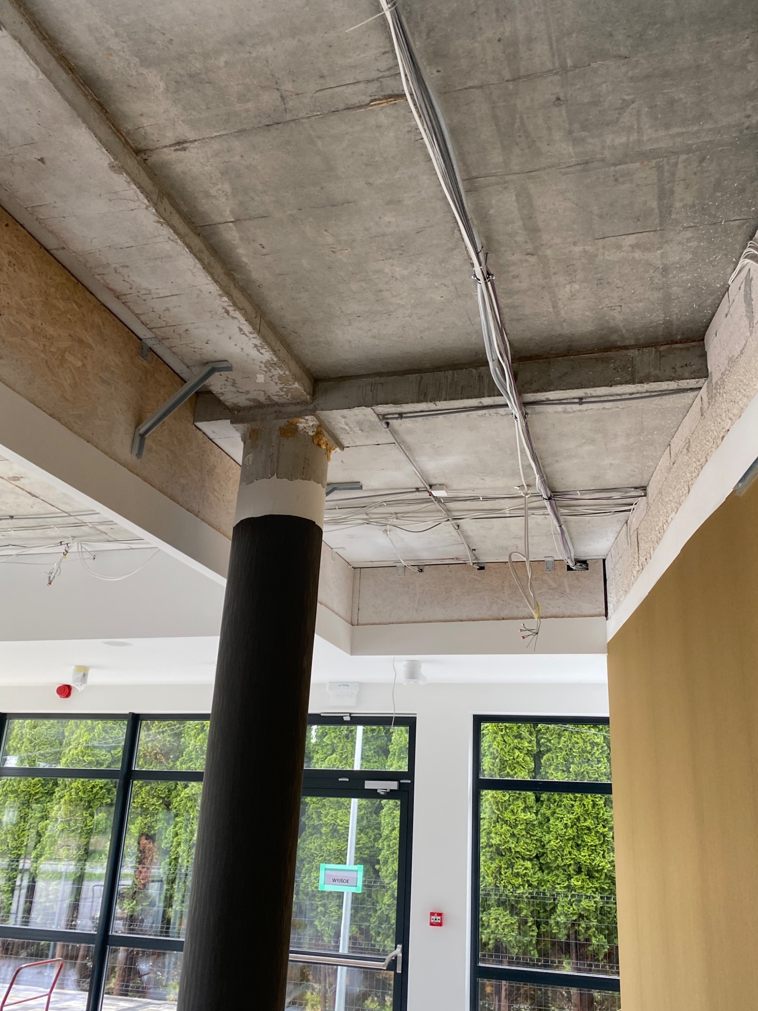 Betonowy strop z widocznymi kablami i rurami, kolumna w trakcie wykończenia, fragment okna z widokiem na zieleń. Etap prac wykończeniowych.