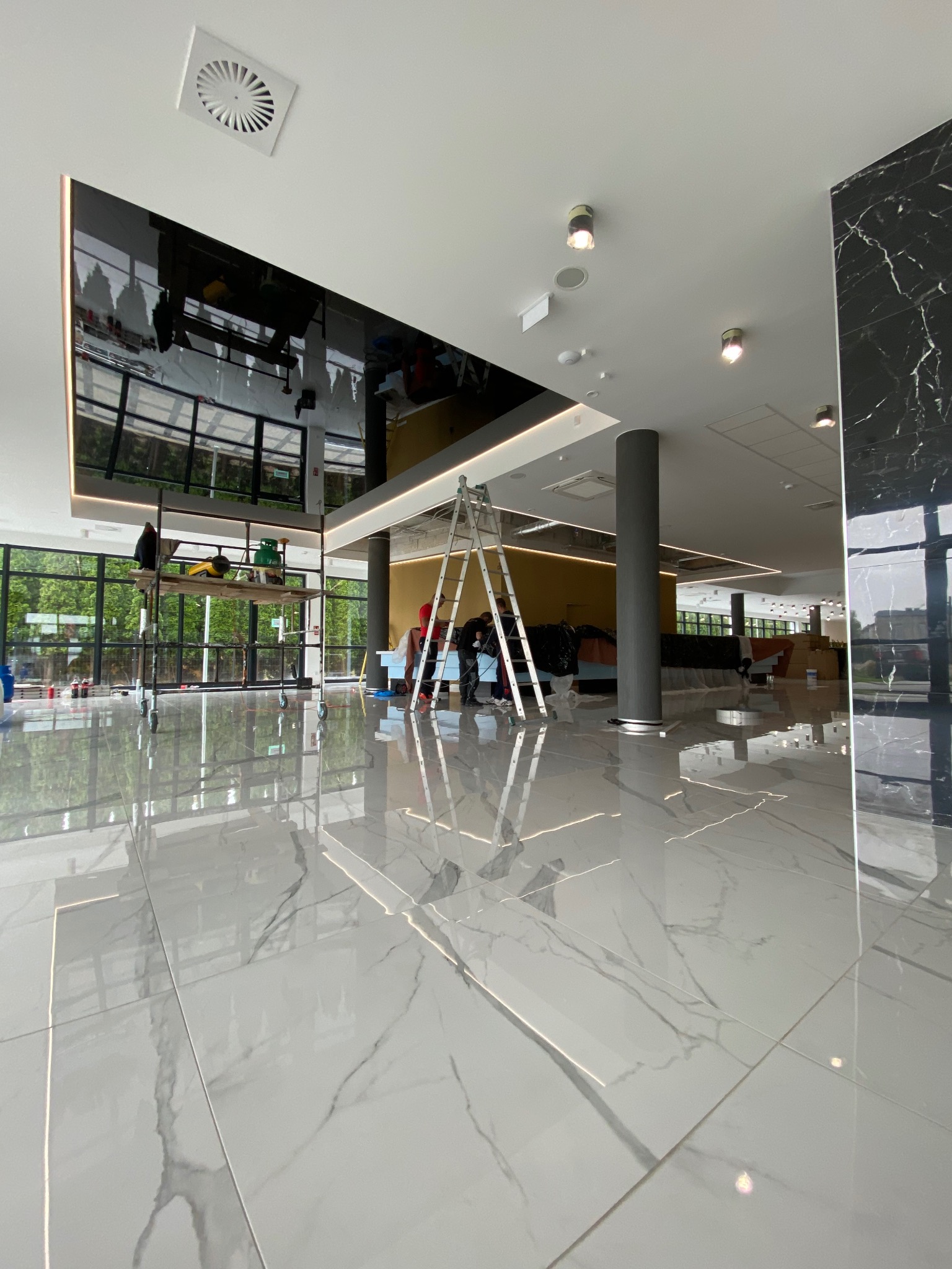 Montaż błyszczącego sufitu napinanego w przestronnym holu z marmurową podłogą. Widoczne rusztowania i drabina, odbicie otoczenia w suficie.