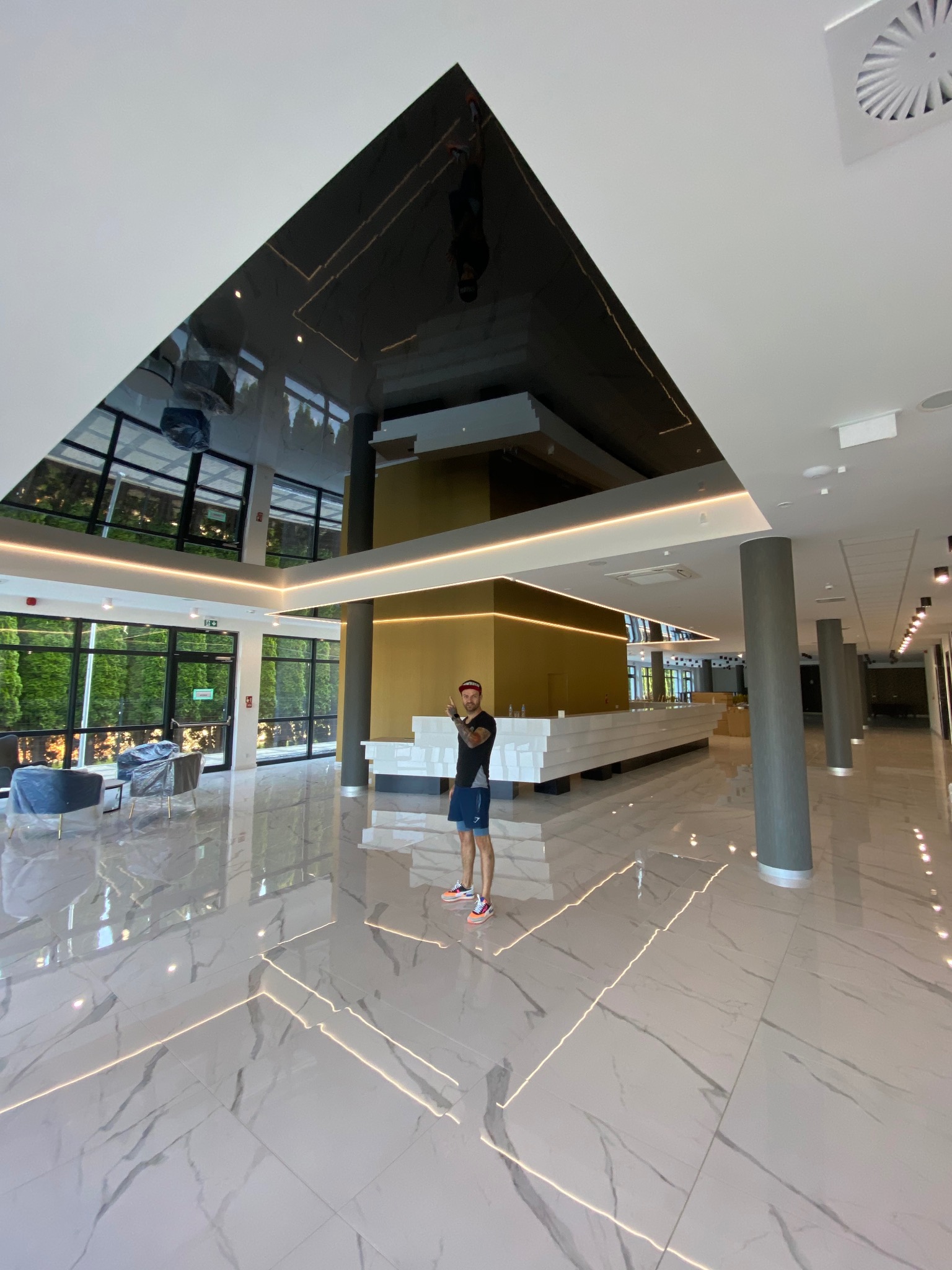 Błyszczący, czarny sufit napinany w przestronnym holu z marmurową podłogą, oświetleniem LED i osobą w tle, odbijający światło i otoczenie.