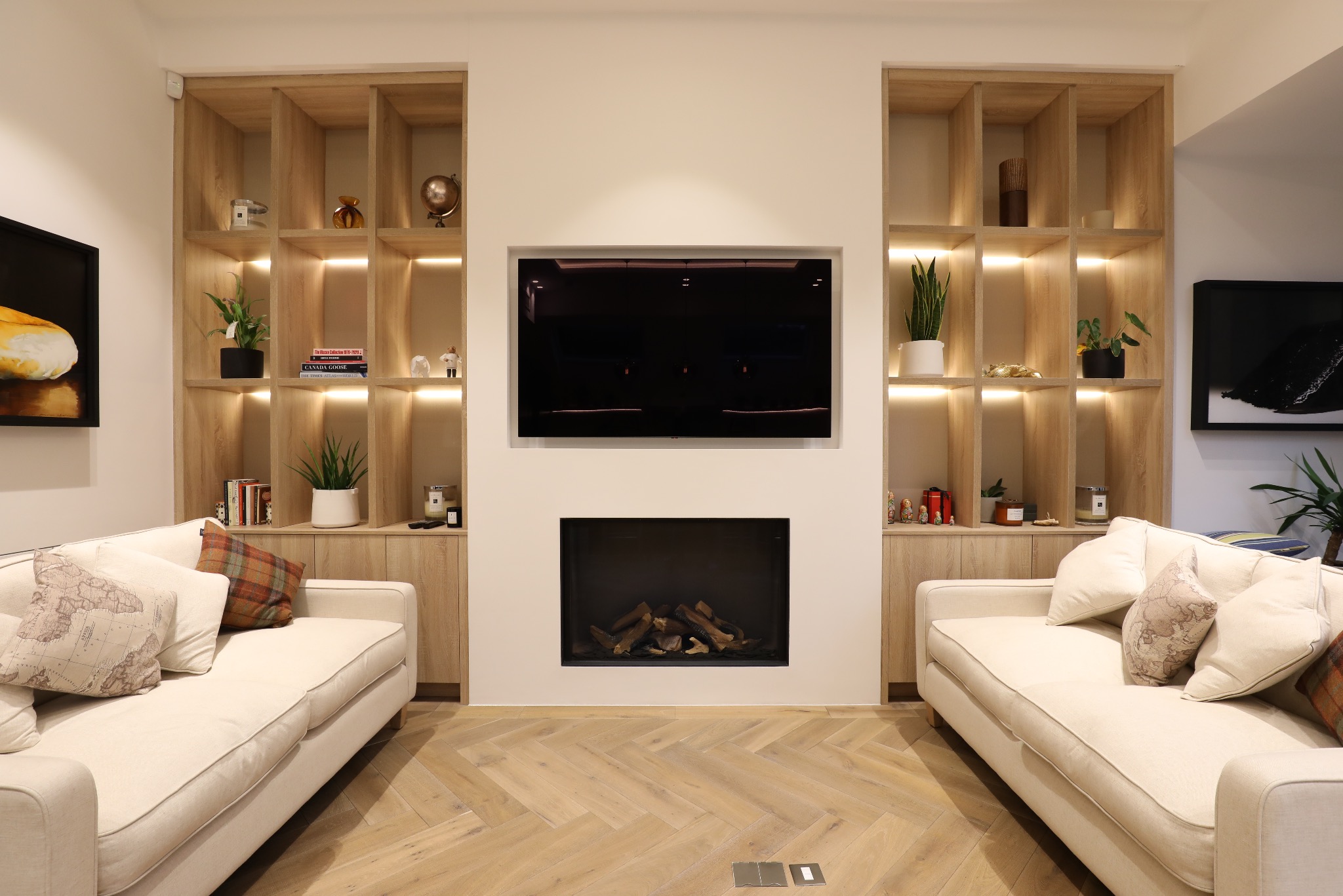 Salon z symetrycznym układem: kremowe sofy, drewniana podłoga w jodełkę, wnęki z półkami po bokach kominka i telewizora. Ciepłe oświetlenie.