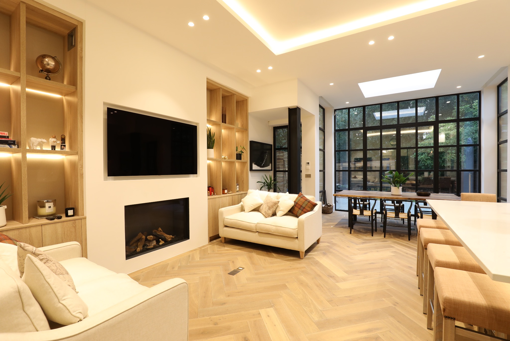 Jasne, przestronne wnętrze salonu z parkietem w jodełkę, kominkiem, zabudową meblową i dużymi oknami. Nowoczesny design z elementami naturalnego drewna i subtelnym oświetleniem.