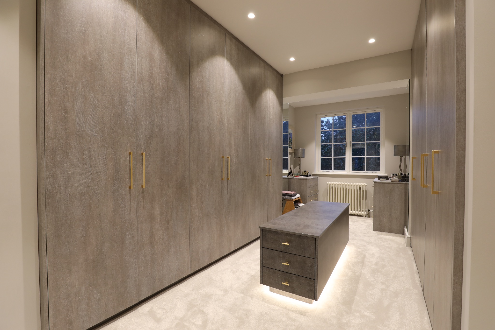 Nowoczesna garderoba z zabudową szaf w odcieniach szarości, z podświetlaną wyspą na środku i złotymi uchwytami, oświetlona punktowo.