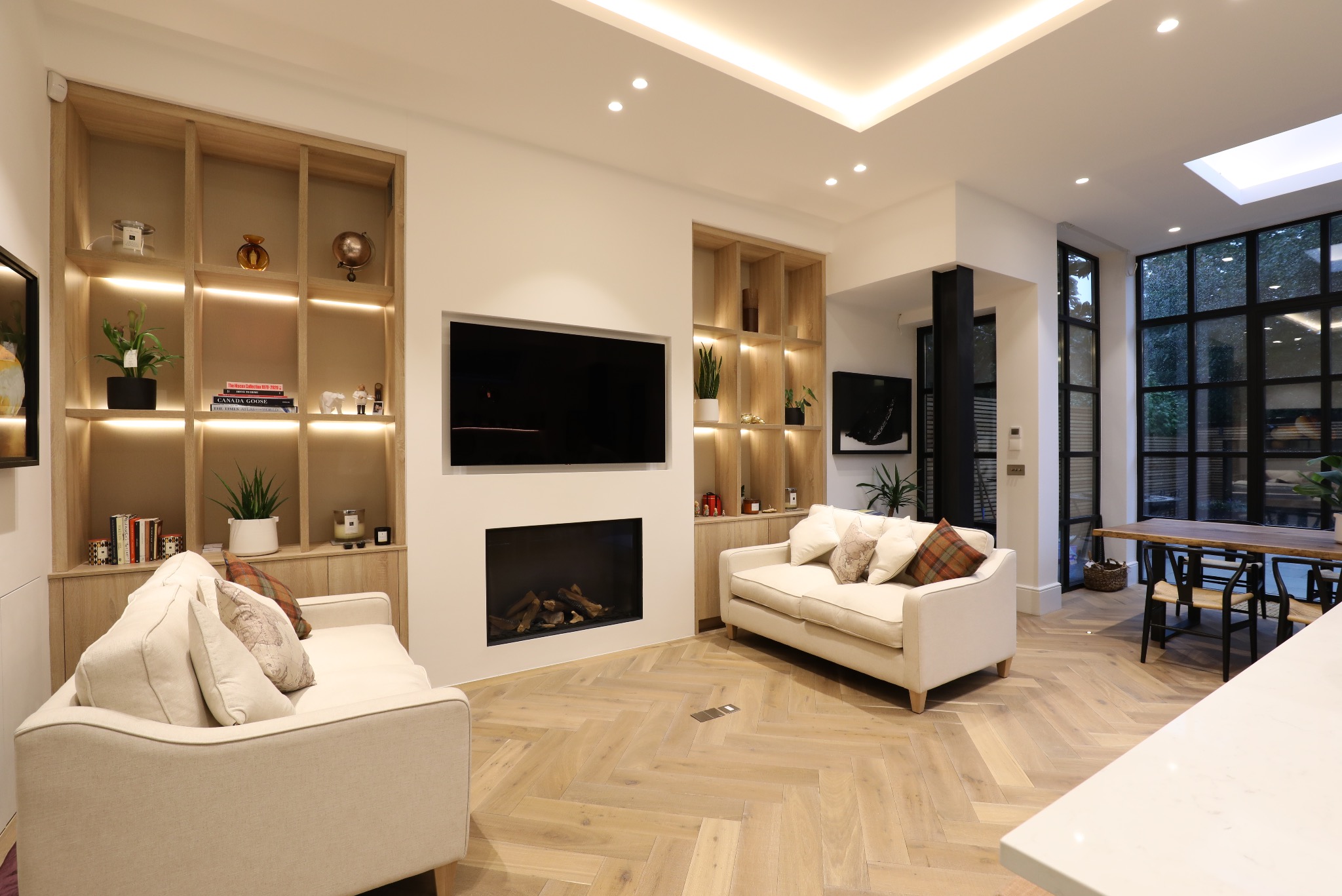 Jasne wnętrze salonu z parkietem w jodełkę, dwiema sofami, kominkiem pod telewizorem i regałami z oświetleniem LED. Duże okna z czarnymi szprosami.