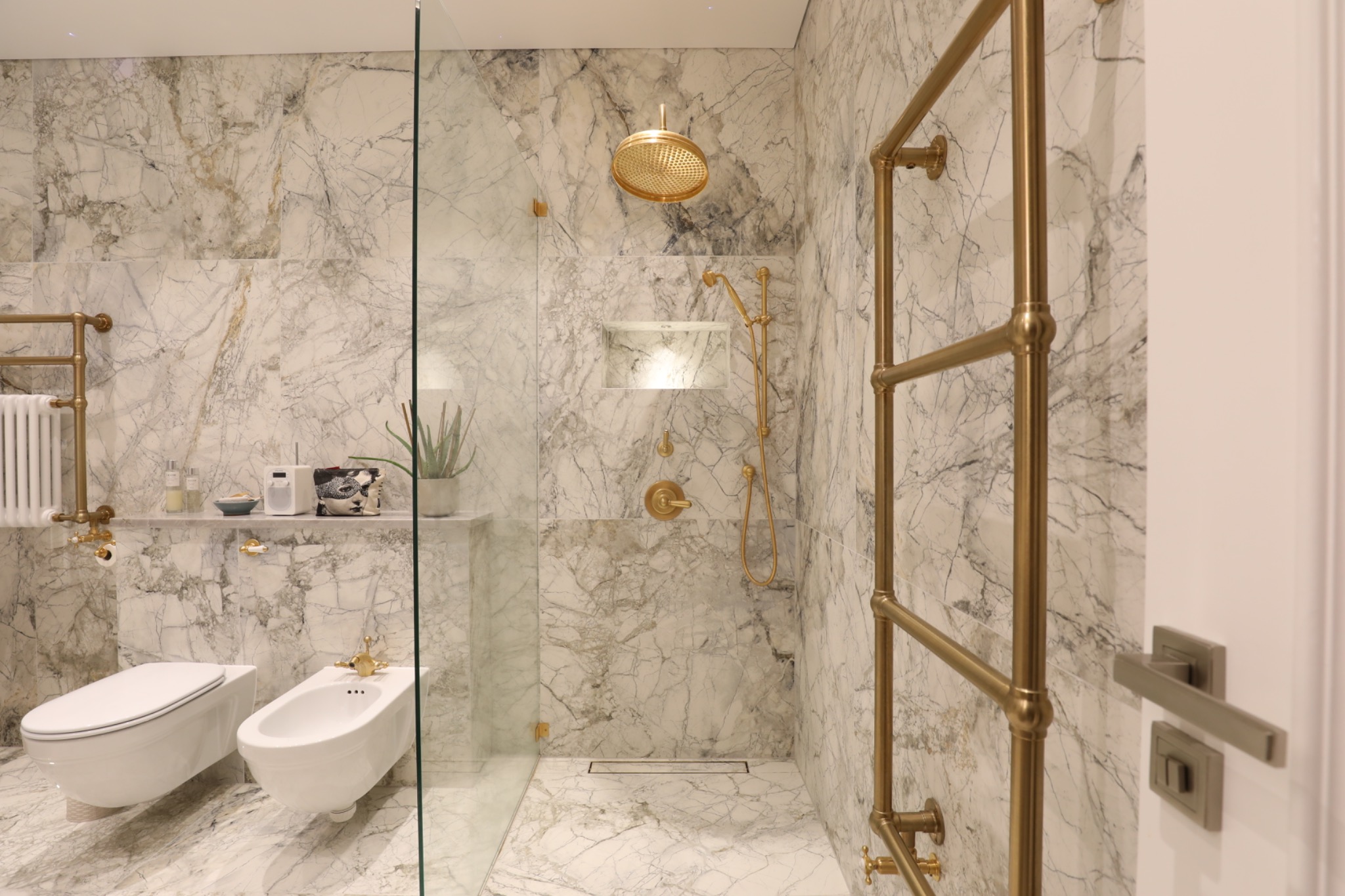 Elegancka łazienka z marmurowymi płytkami, złotą armaturą i szklaną kabiną prysznicową. Widoczny grzejnik drabinkowy i toaleta z bidetem.
