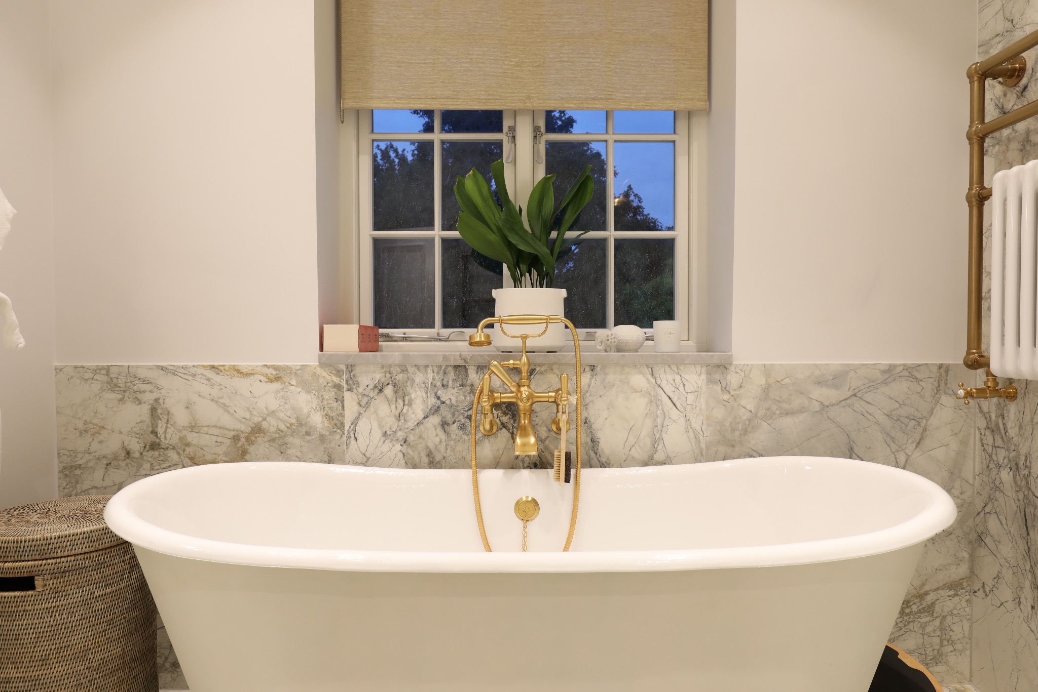 Elegancka, wolnostojąca wanna na tle marmurowej ściany. Złota armatura, roślina w doniczce na parapecie. Luksusowa łazienka po remoncie, detale wykończeniowe.