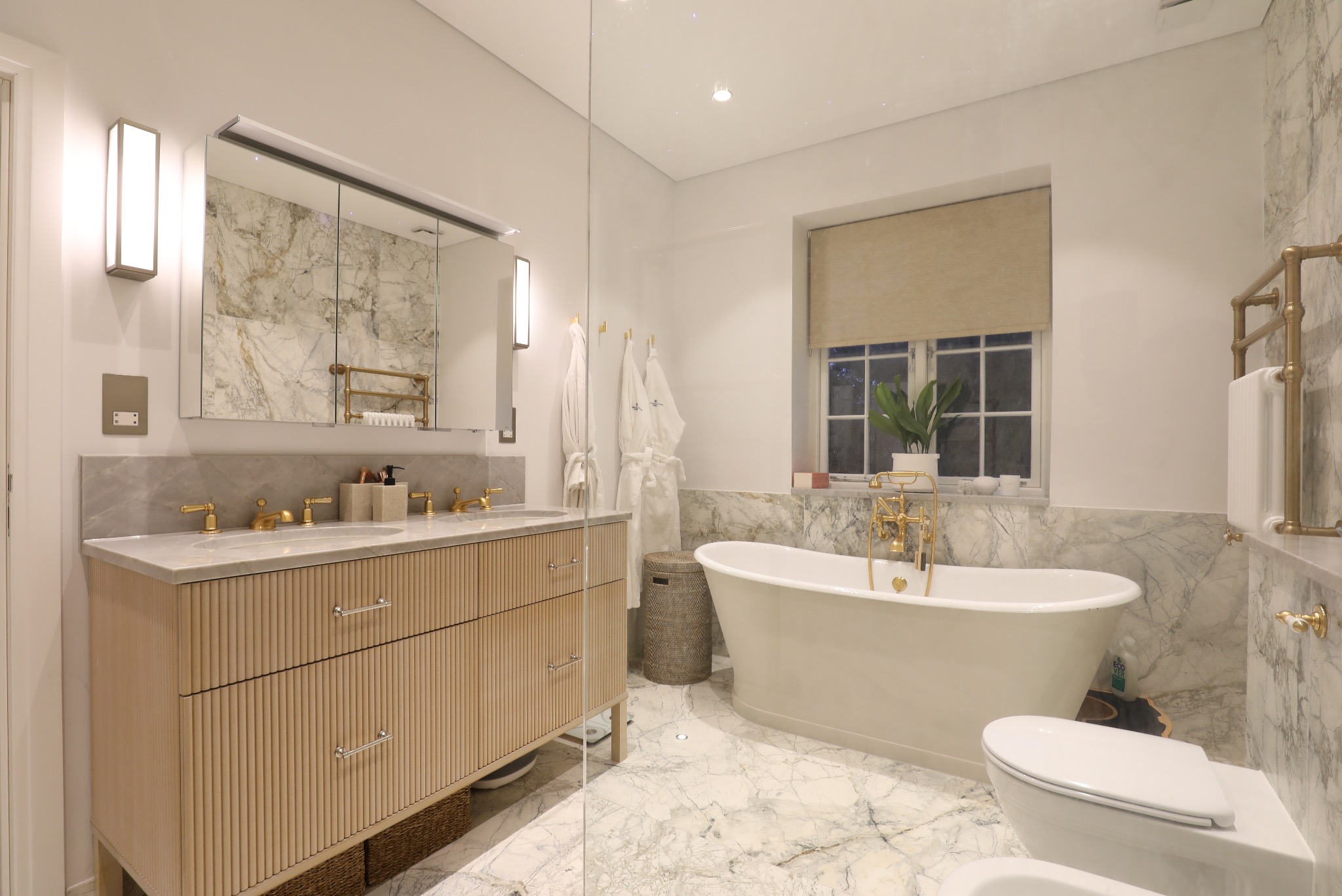 Elegancka łazienka z marmurowymi ścianami i podłogą, wolnostojąca wanna z złotą armaturą, drewniana szafka pod umywalkę z ryflowanymi frontami i lustro z oświetleniem.