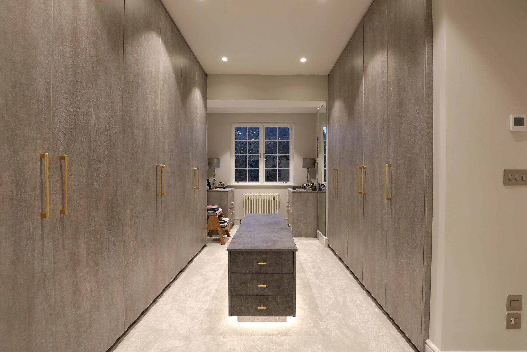 Nowoczesna garderoba z szarymi szafami wnękowymi i złotymi uchwytami, oświetlona punktowo, z wyspą na środku i oknem w tle. Eleganckie wykończenie.