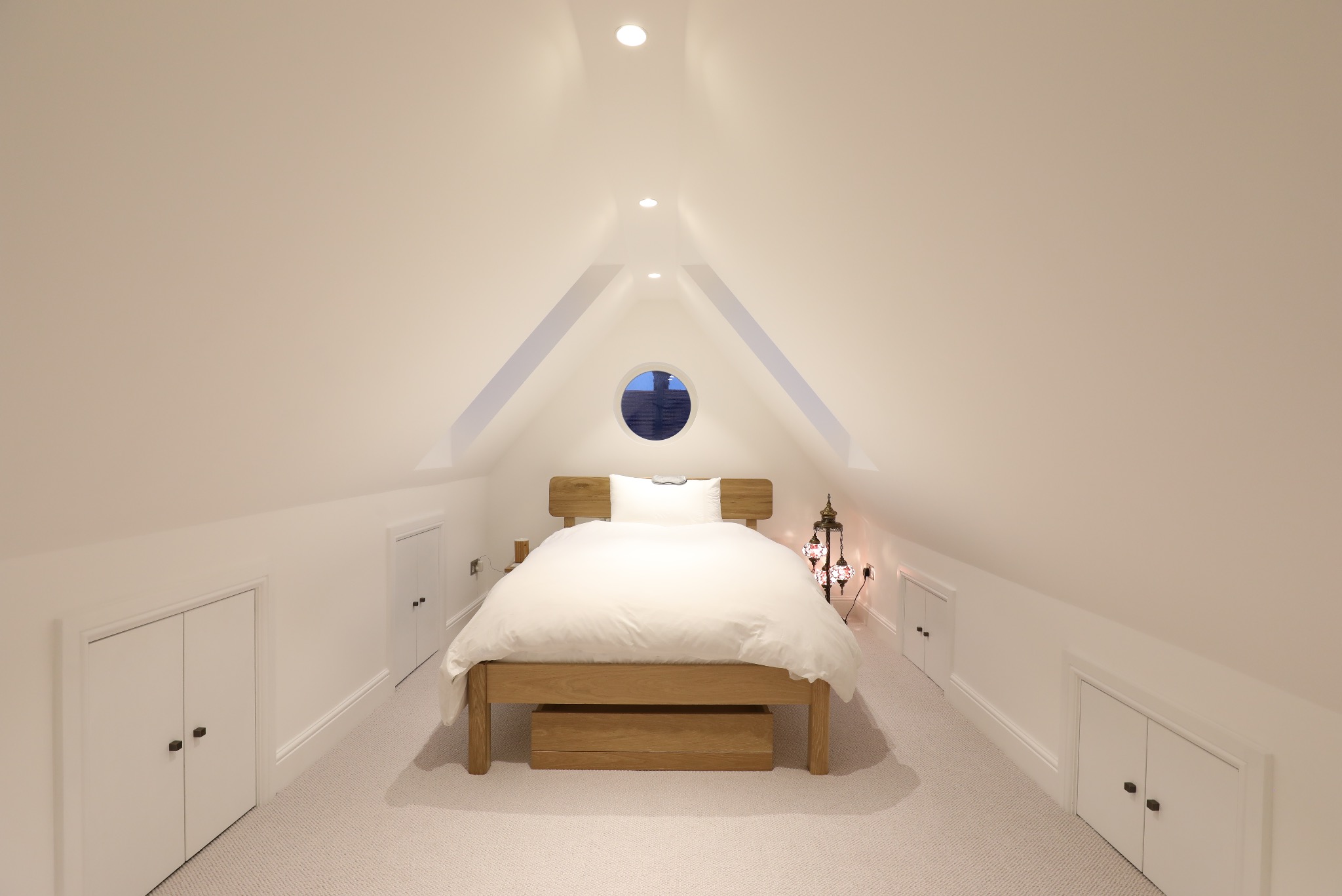 Jasna sypialnia na poddaszu z drewnianym łóżkiem, okrągłym oknem i wbudowanymi szafkami. Minimalistyczny design z orientalną lampą.