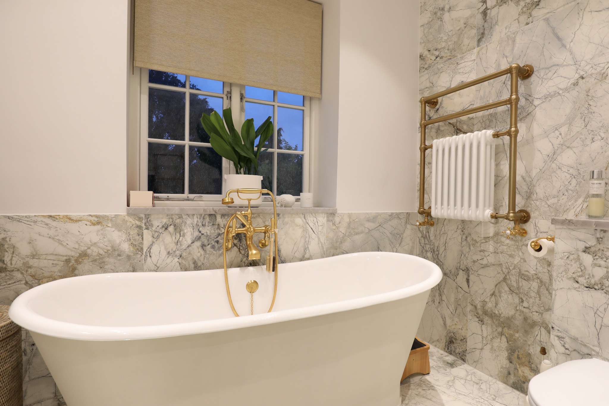 Elegancka łazienka z wolnostojącą wanną, złotą armaturą i marmurowymi płytkami. Grzejnik drabinkowy w kolorze złota. Roślina w oknie.
