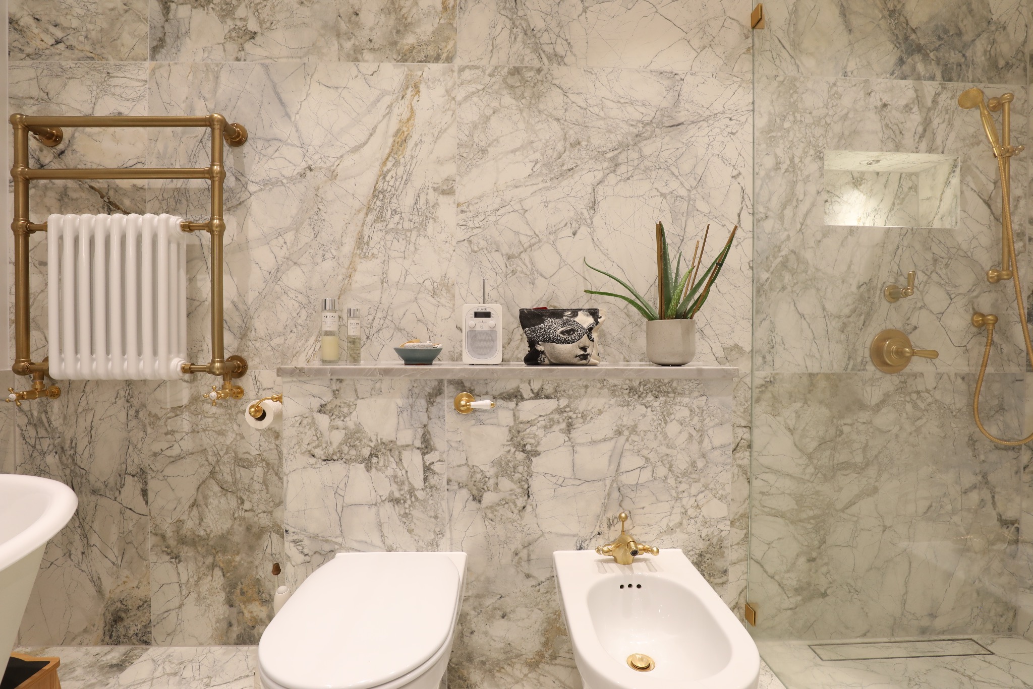 Elegancka łazienka z marmurowymi płytkami, złotą armaturą i grzejnikiem drabinkowym. Na półce dekoracje: radio, roślina i kosmetyki. Luksusowe wykończenie.