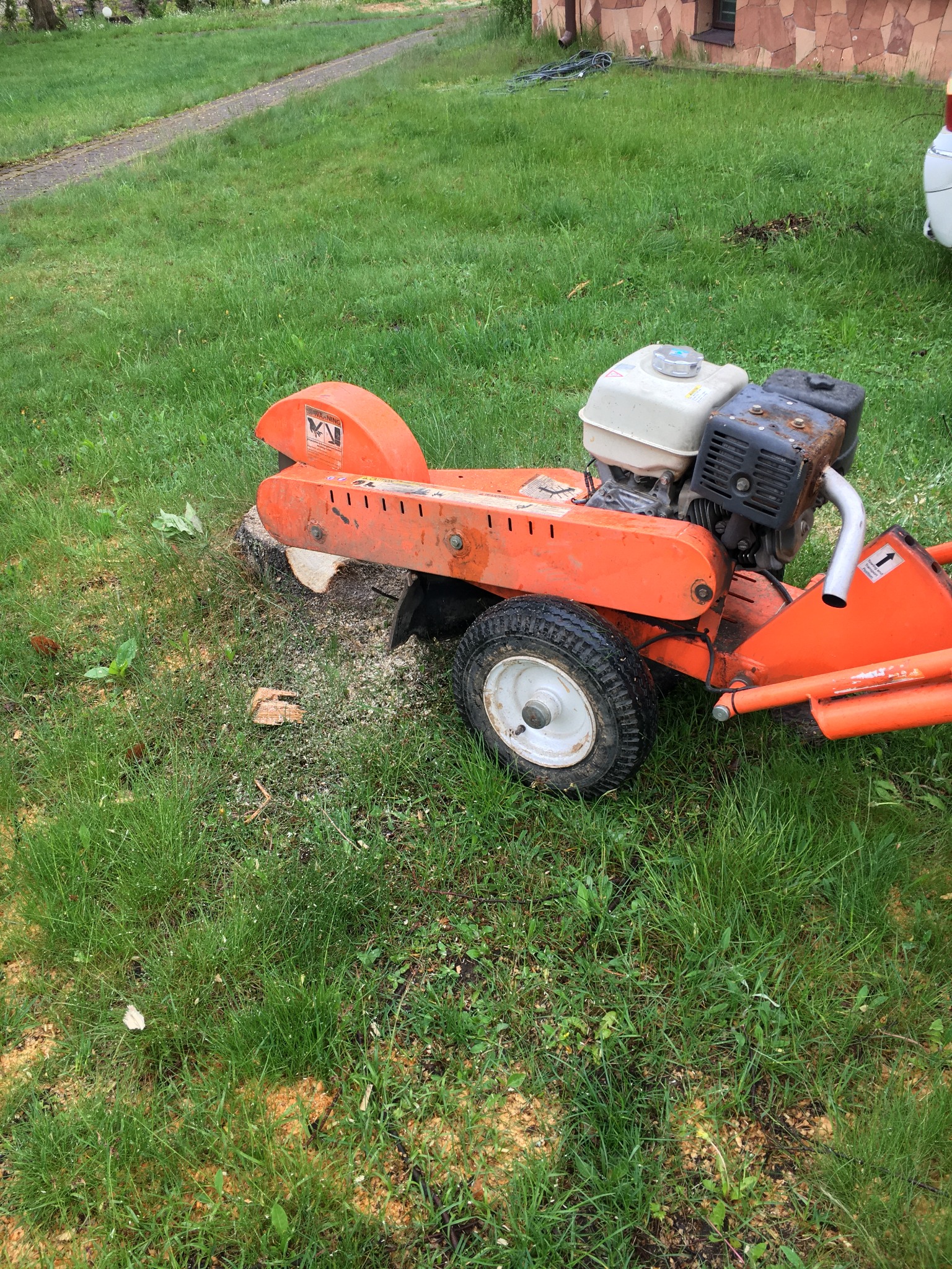 Pomarańczowa frezarka do pni drzew na trawniku po usunięciu pnia, z widocznymi wiórami i resztkami drewna w trawie.