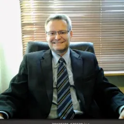 Uśmiechnięty mężczyzna w garniturze i okularach siedzi na krześle biurowym na tle żaluzji okiennych.