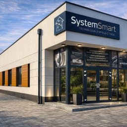 SystemSmart Łukasz Waśniewski - Nowoczesny budynek firmy SystemSmart z logo i hasłem 'Technologia z myślą o Tobie'. Widoczne okna z reklamami partnerów i zadbany chodnik.