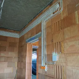 Surowe wnętrze z ceglanymi ścianami w trakcie prac. Widoczne ułożone wzdłuż sufitu i ściany przewody elektryczne, puszka instalacyjna i fragment otworu drzwiowego zabezpieczonego folią.