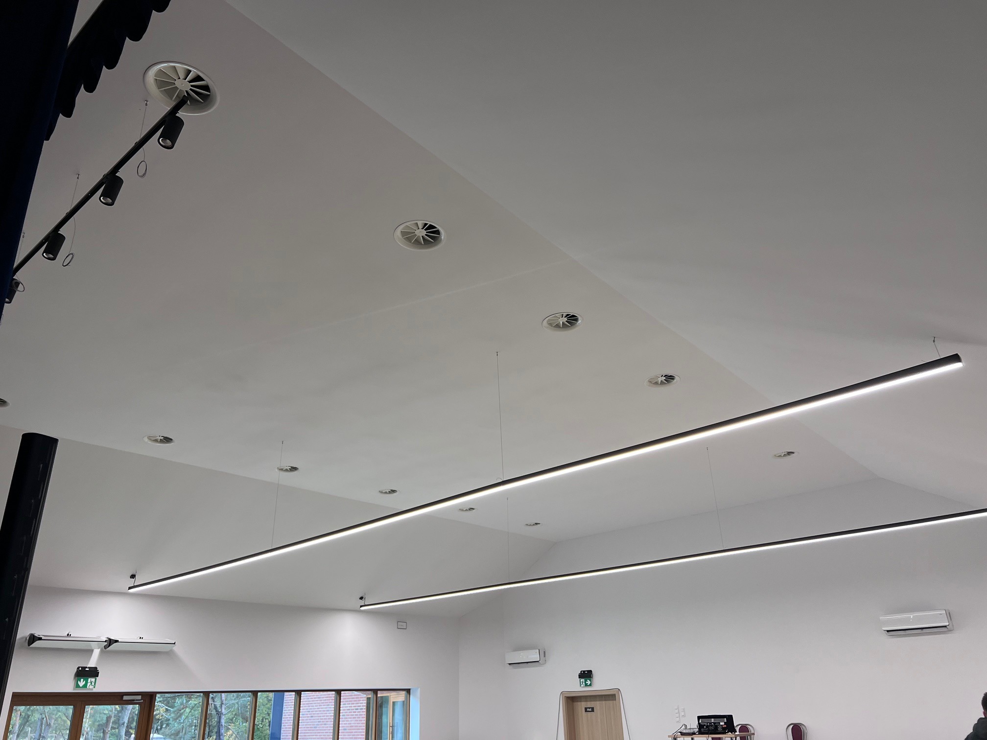 Nowoczesne oświetlenie LED w przestronnym wnętrzu z białym sufitem, widoczne lampy liniowe, wentylacja sufitowa i elementy instalacji elektrycznej.