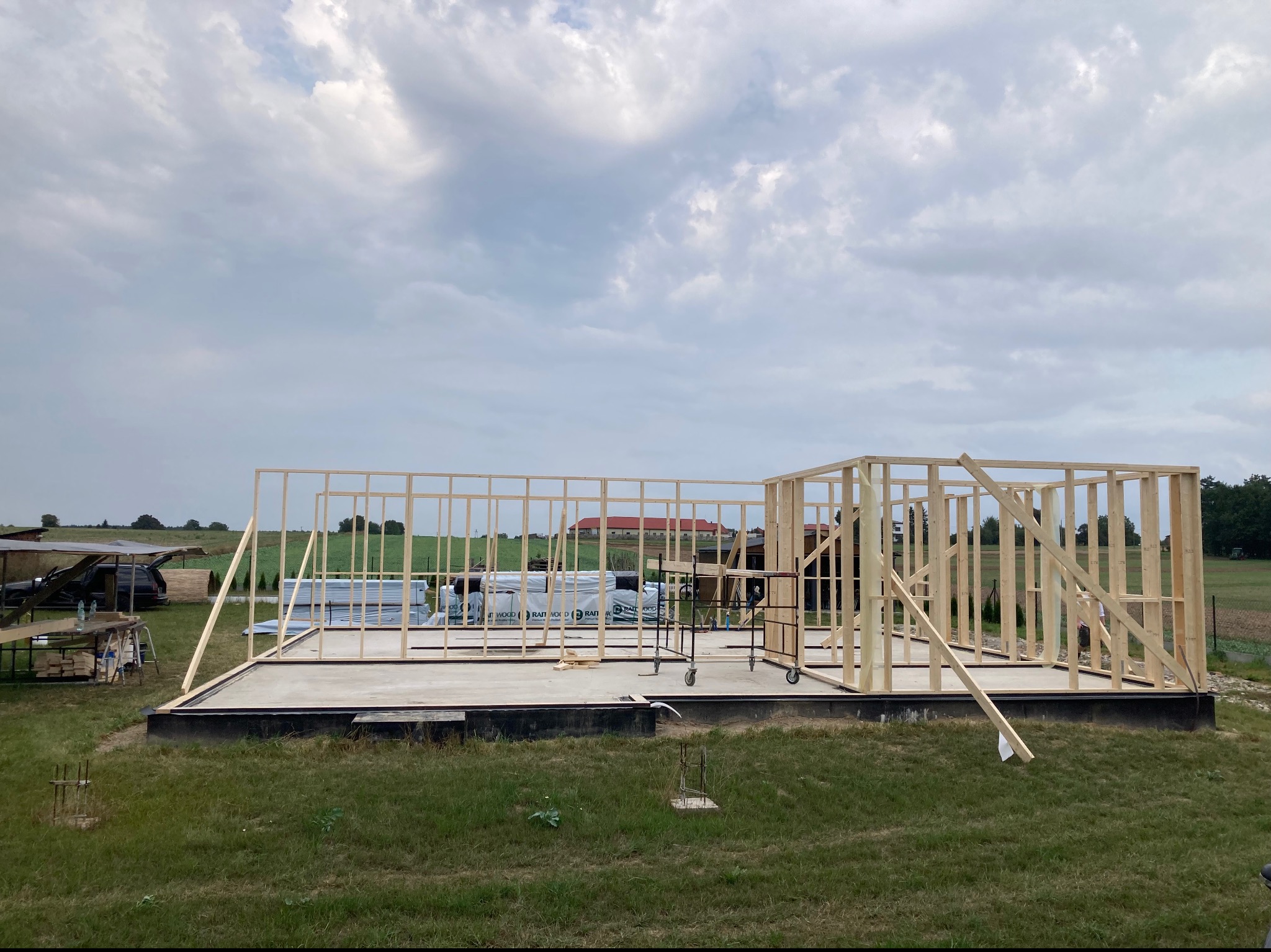 Wznoszenie drewnianej konstrukcji szkieletowej domu na betonowej płycie fundamentowej, widoczne elementy konstrukcyjne, rusztowanie na kółkach i materiały budowlane w tle, pochmurne niebo.