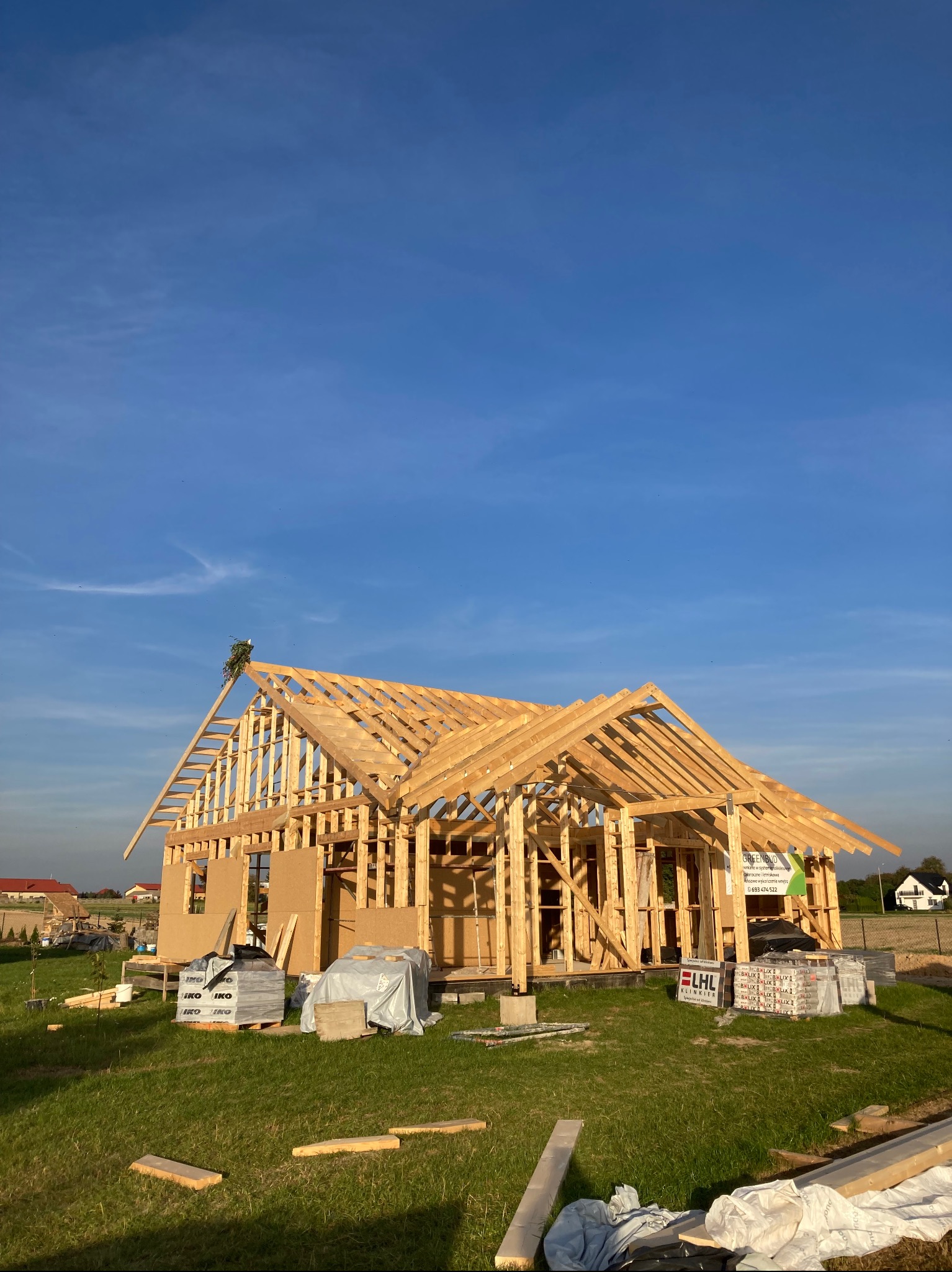 Wznoszona konstrukcja drewniana domu jednorodzinnego w stanie surowym otwartym, z widocznym odeskowaniem dachu, belkami stropowymi i ścianami, na tle błękitnego nieba i zielonej trawy. Na szczycie...