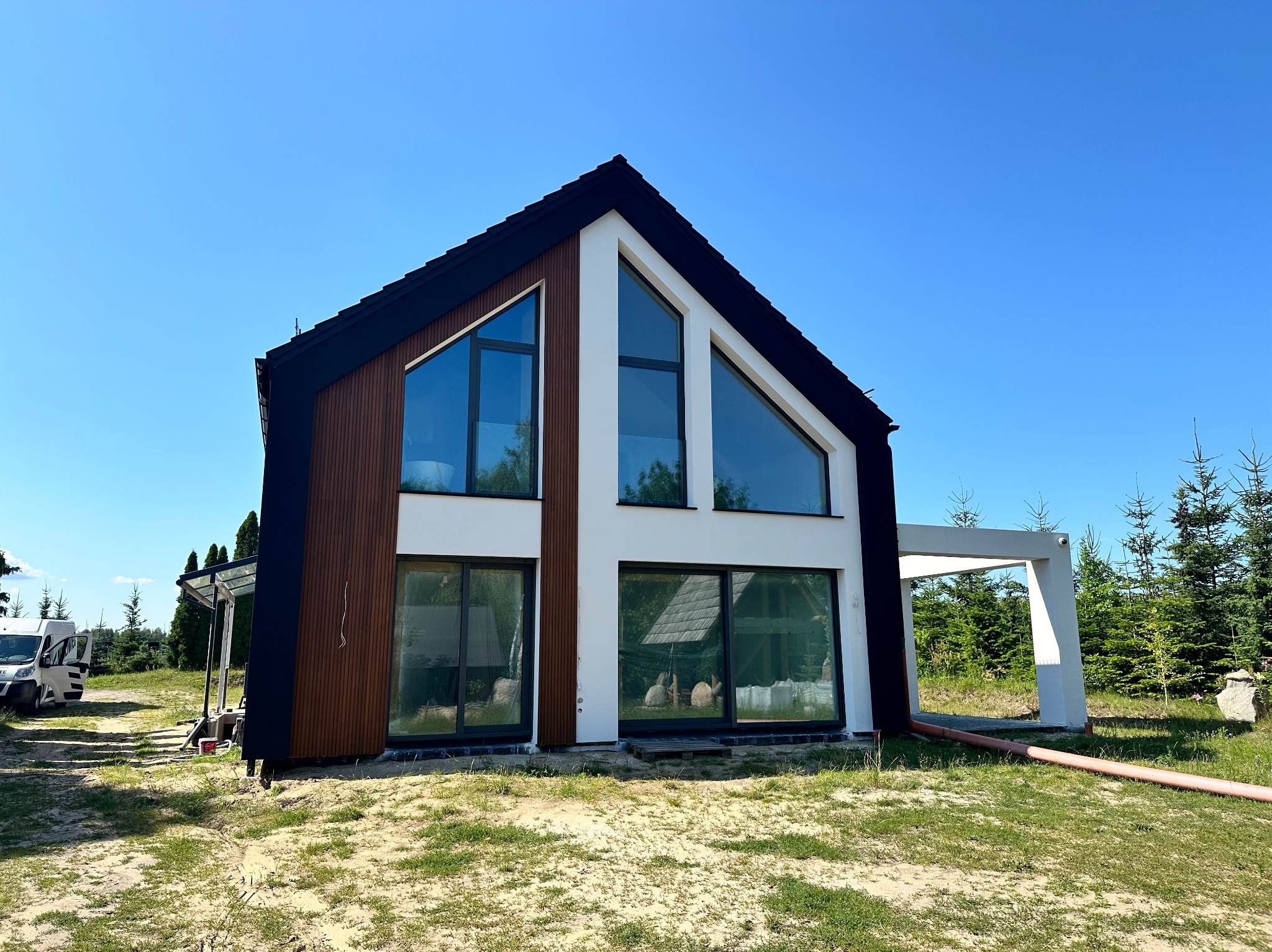 Nowoczesny dom jednorodzinny z elementami drewnianej elewacji, dużymi oknami i czarnym dachem w słoneczny dzień. Widoczny fragment terenu wokół budynku z piaskiem i trawą.