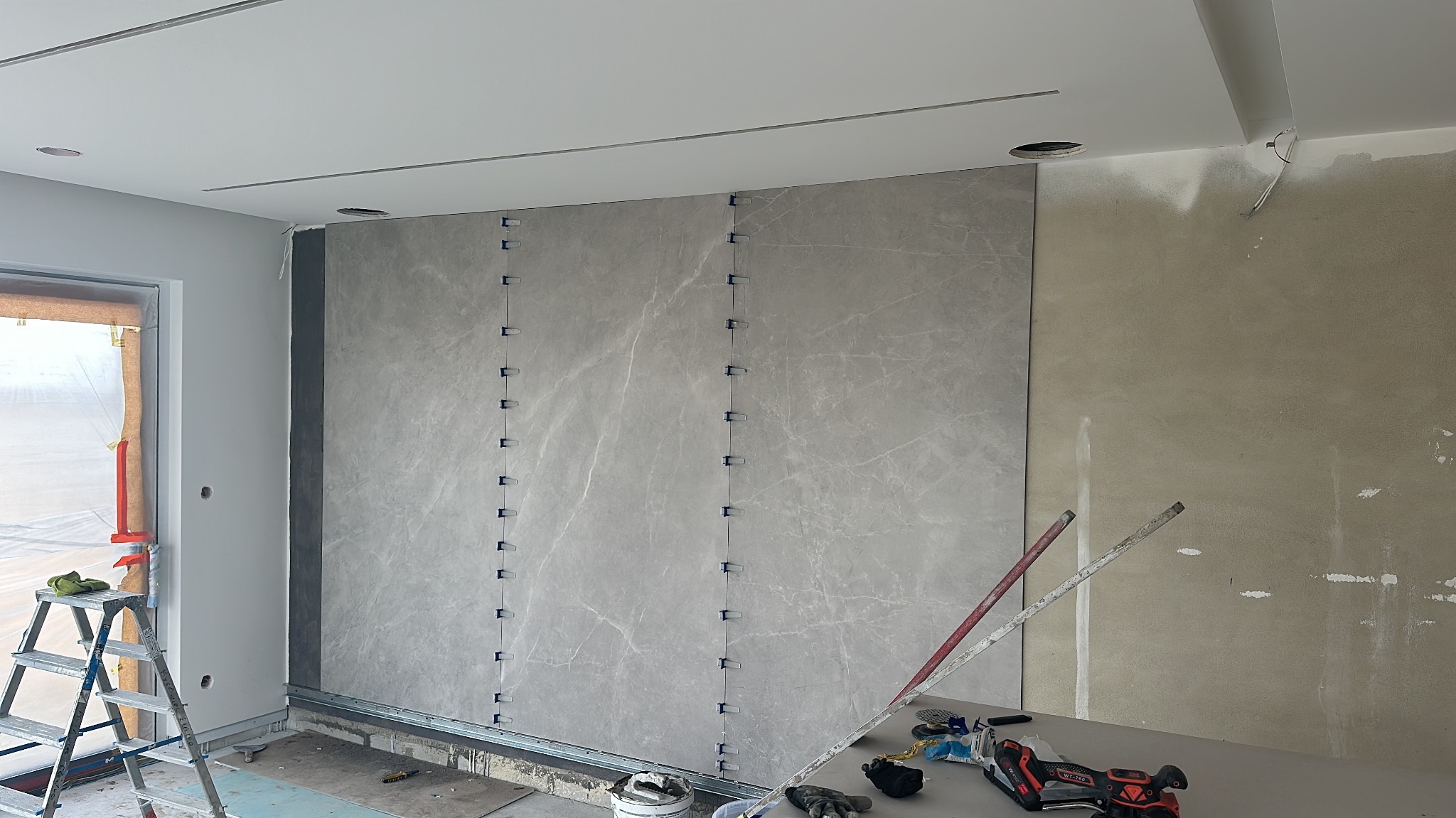 Montaż wielkoformatowych płyt na ścianie, widoczne listwy startowe i system poziomowania płytek, drabina i narzędzia w tle.