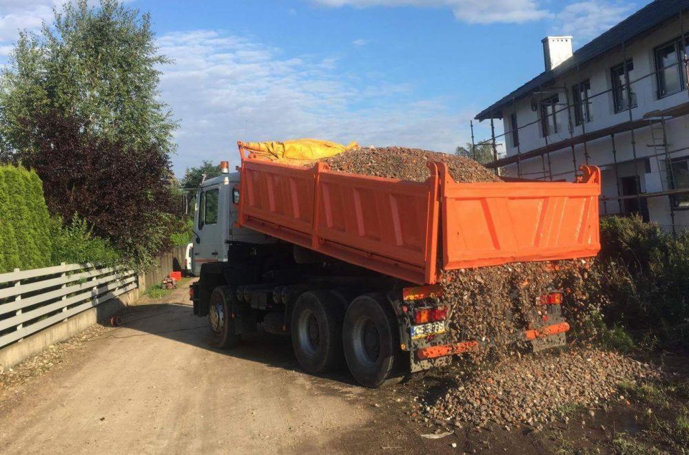 Pomarańczowa wywrotka wysypuje gruz na ziemię obok budynku w trakcie prac budowlanych, widoczne rusztowania.