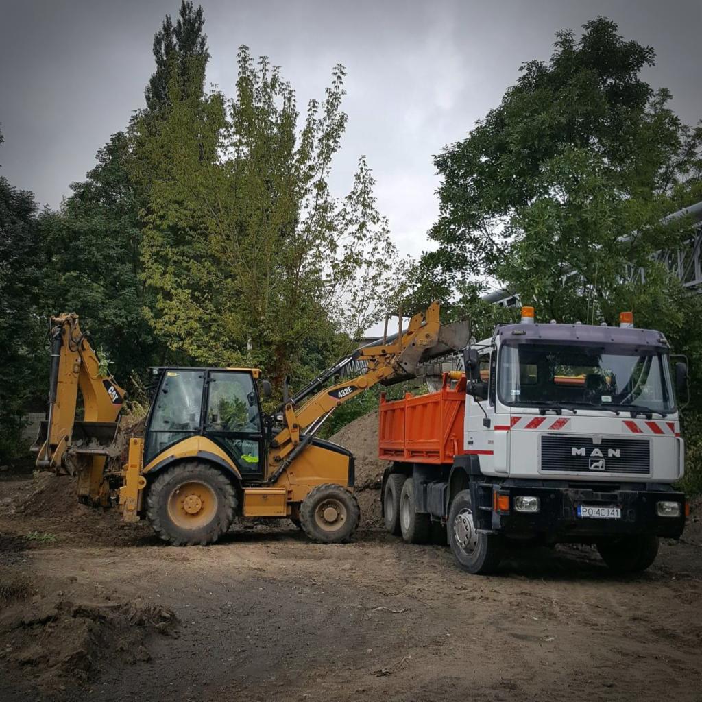Żółta koparko-ładowarka CAT 432E nabiera ziemię i ładuje ją do pomarańczowej skrzyni wywrotki MAN na tle drzew i pochmurnego nieba.