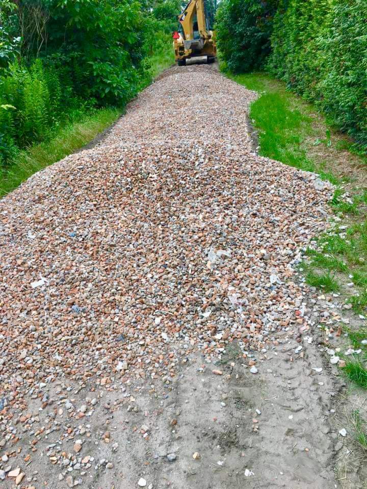 Żwirowa droga usypana kopcem, w tle żółta koparka na tle zieleni.