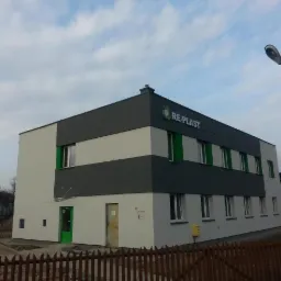 Dwukondygnacyjny budynek biurowy z szarą i białą elewacją w Oświęcimiu, z widocznym logo firmy RE-PLAST na górnej części fasady, zielone obramowania okien i drzwi, oświetlony latarnią uliczną.