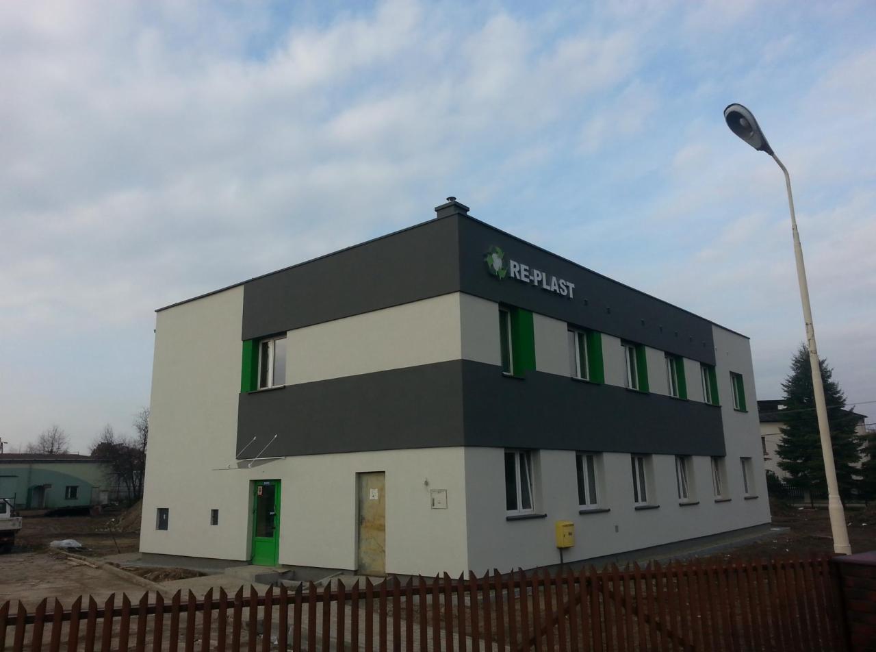 Dwukondygnacyjny budynek biurowy z szarą i białą elewacją w Oświęcimiu, z widocznym logo firmy RE-PLAST na górnej części fasady, zielone obramowania okien i drzwi, oświetlony latarnią uliczną.
