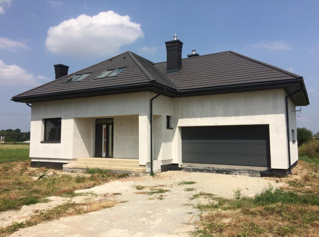 Surowy, jednokondygnacyjny dom z szarą dachówką i bramą garażową, w trakcie wykończenia elewacji zewnętrznej, widoczny fragment instalacji odprowadzającej wodę deszczową.