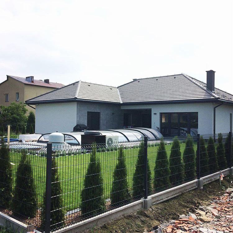 Nowoczesny dom jednorodzinny z szarą dachówką, widok zza metalowego ogrodzenia z betonową podmurówką i rzędem małych, gęsto posadzonych iglaków; w tle zadaszony basen i klimatyzator zewnętrzny.
