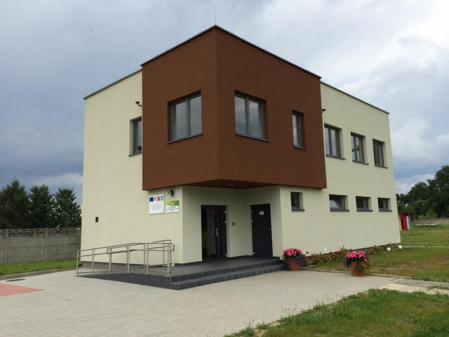 Dwukondygnacyjny budynek o nowoczesnej architekturze z brązowym akcentem na elewacji, rampą dla niepełnosprawnych przy wejściu i kwiatami w donicach.