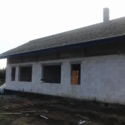 Niedokończony budynek z szarym dachem i kominem, widoczne pustaki, brak okien, drabina i gruz na ziemi.